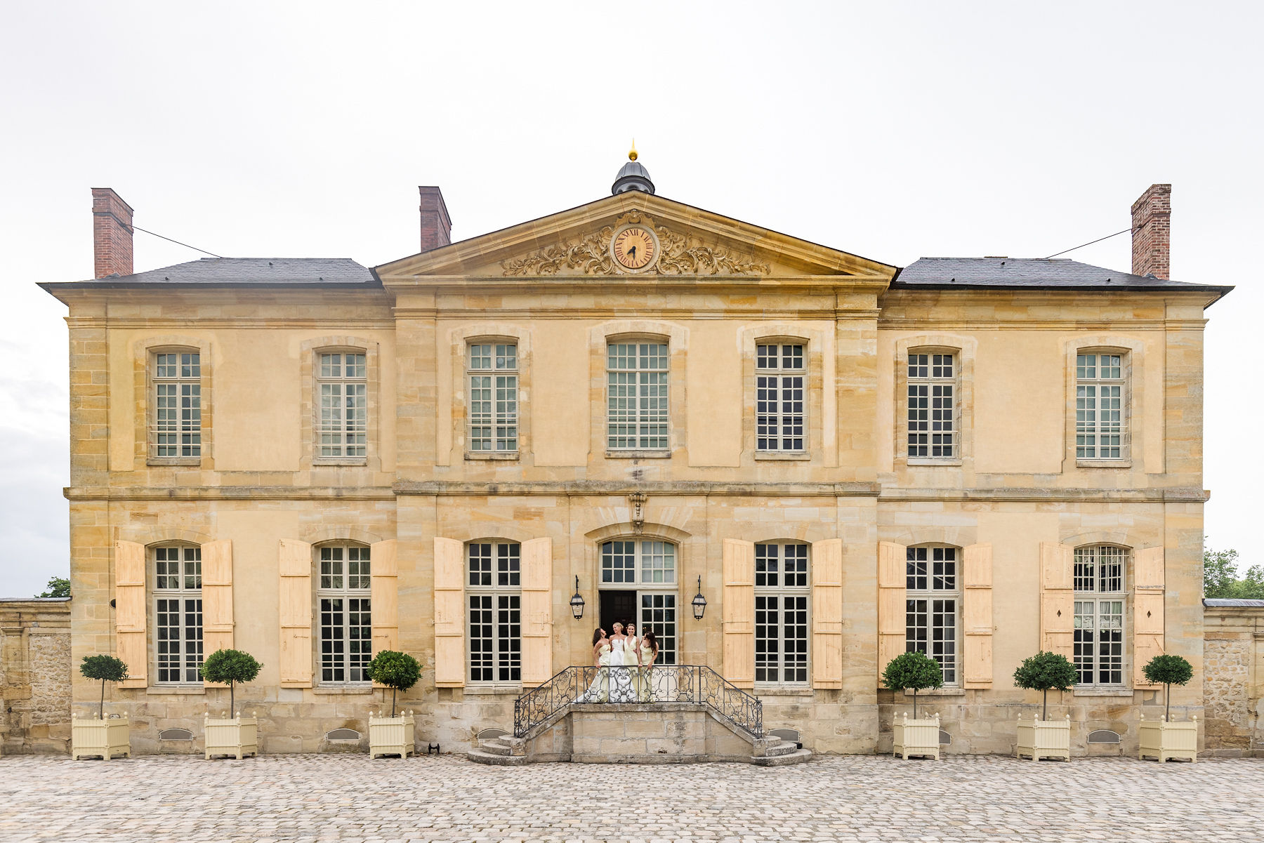 Bride and groom on wrought-iron balcony of symmetrical classical stone manor with gilded pediment and topiary courtyard