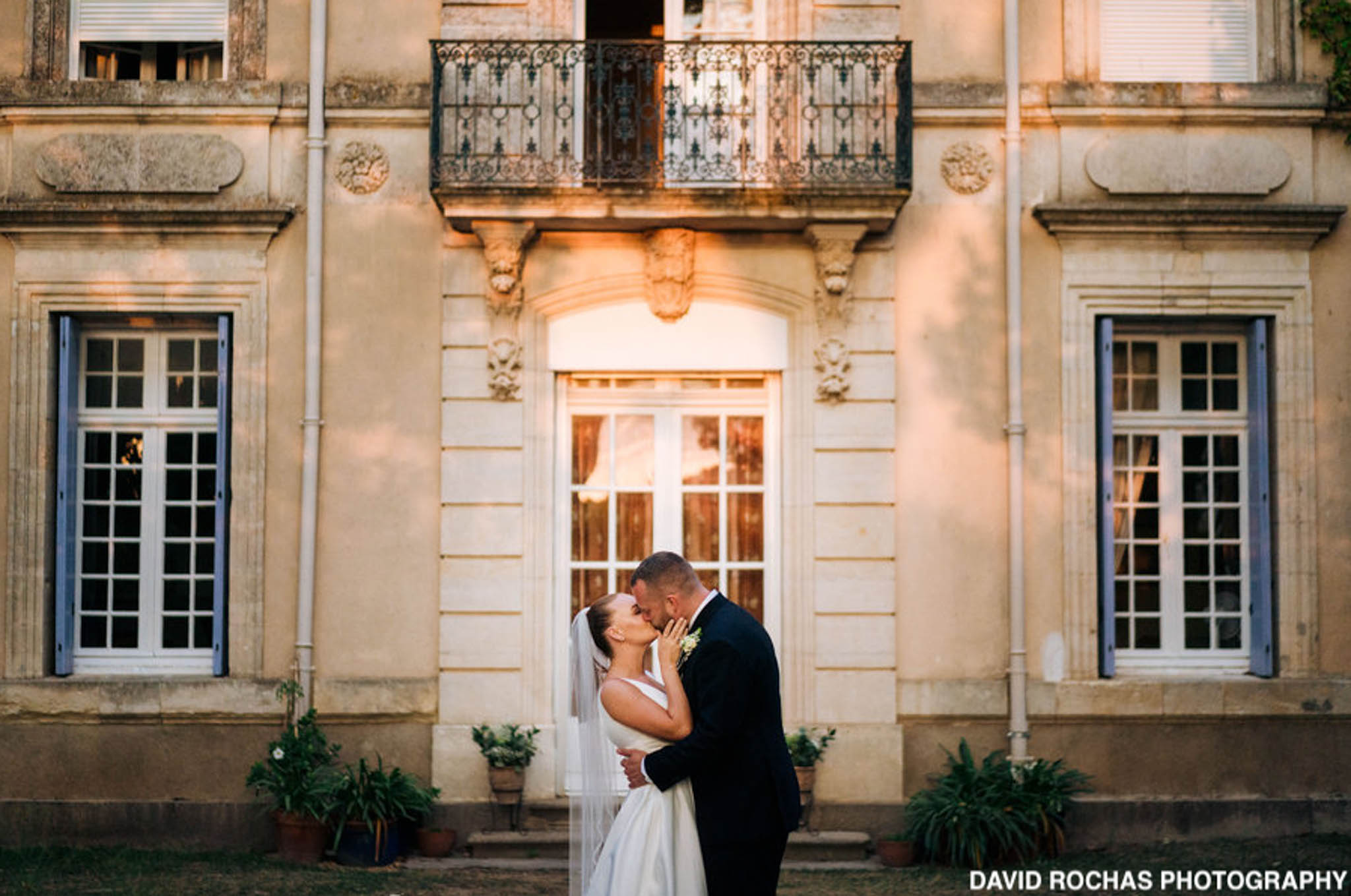 Bride and groom kissing beneath ornate arched doorway of limestone chateau facade in golden hour light
