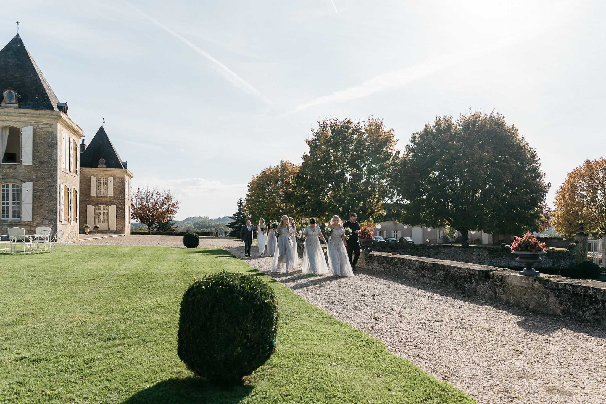 Bride and powder blue bridesmaids walking gravel path toward slate-roofed chateau with topiary gardens