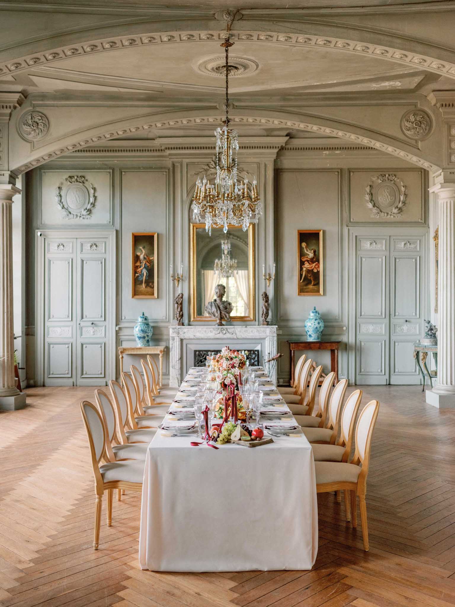 A styled wedding reception tablescape set inside a grand French château ballroom or formal dining room. A long rectangular table covered in a white linen tablecloth seats approximately 14 guests, fitted with gold-framed Louis XVI-style chairs upholstered in cream fabric. The centerpiece features a lush arrangement of blush, coral, and peach blooms alongside deep red taper candles, loose ribbon in burgundy, and a spread of fresh fruit including grapes, pears, and an apple on a wooden board. Place settings include white plates, multiple crystal glasses, and gold flatware. The room features pale sage-green painted wood paneling with ornate plasterwork medallions and cornicing, a crystal and brass chandelier overhead, a marble fireplace flanked by classical sculptures and blue-and-white ceramic vases, and framed figurative oil paintings in gilded frames. The floor is herringbone parquet wood. The overall styling palette combines ivory, gold, sage green, and deep burgundy accents in a classic French interior style. Wide shot taken from a slightly elevated angle looking down the length of the table toward the fireplace. Potential venue feature image.
