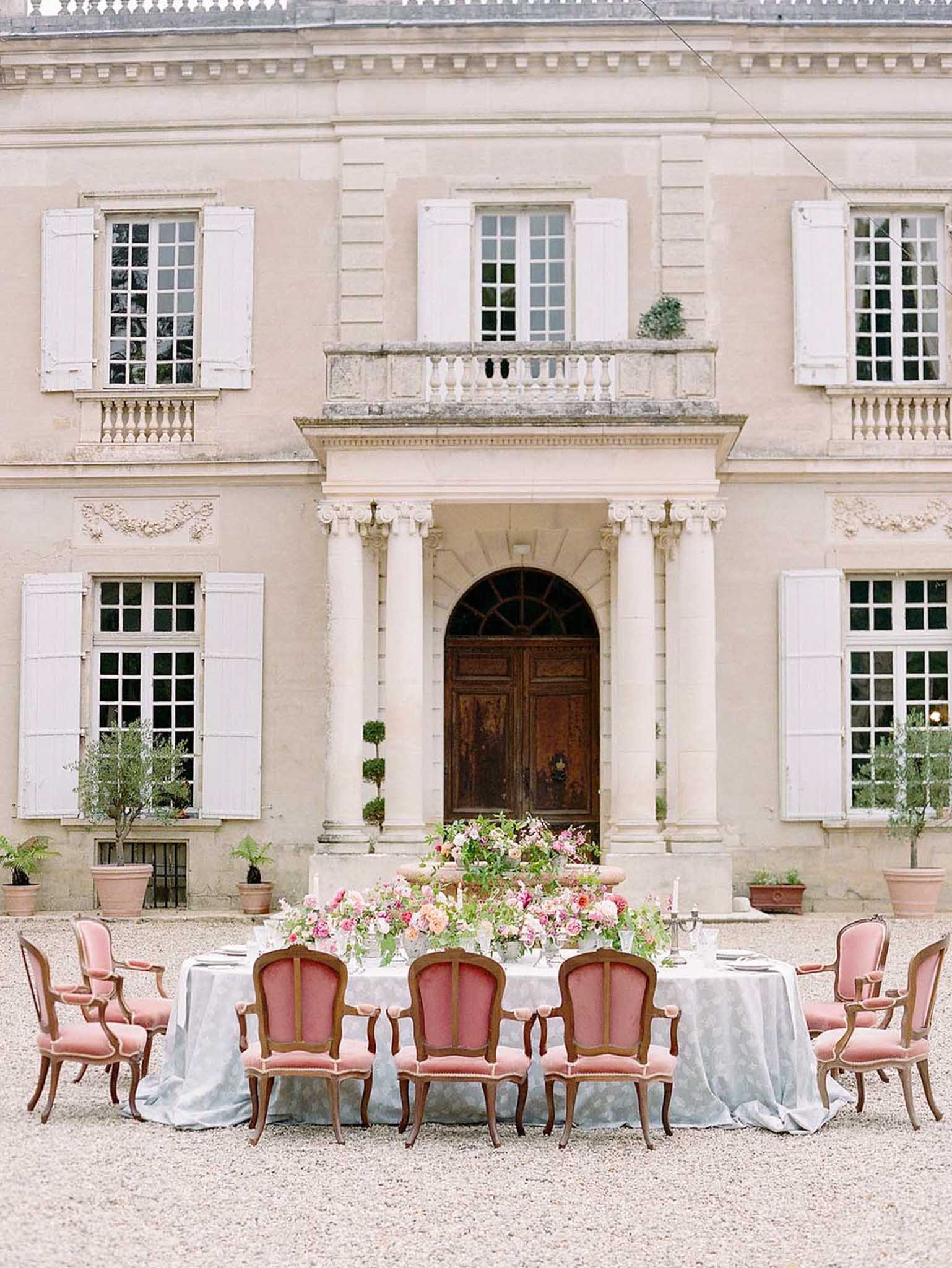 Oval dining table with dusty rose velvet chairs and coral rose centerpiece before a classical French chateau entrance