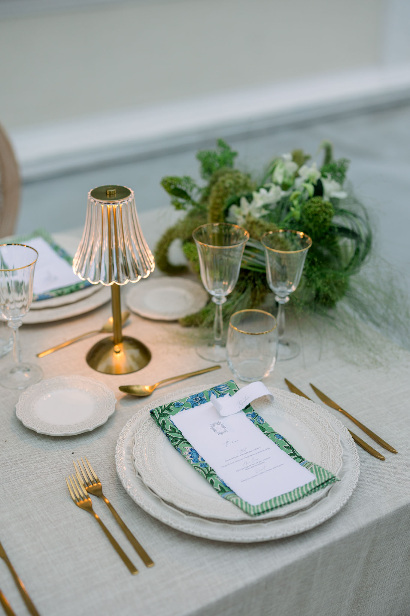 Table setting with white textured charger plates, gold flatware, botanical menu ribbon, and brass lamp centerpiece