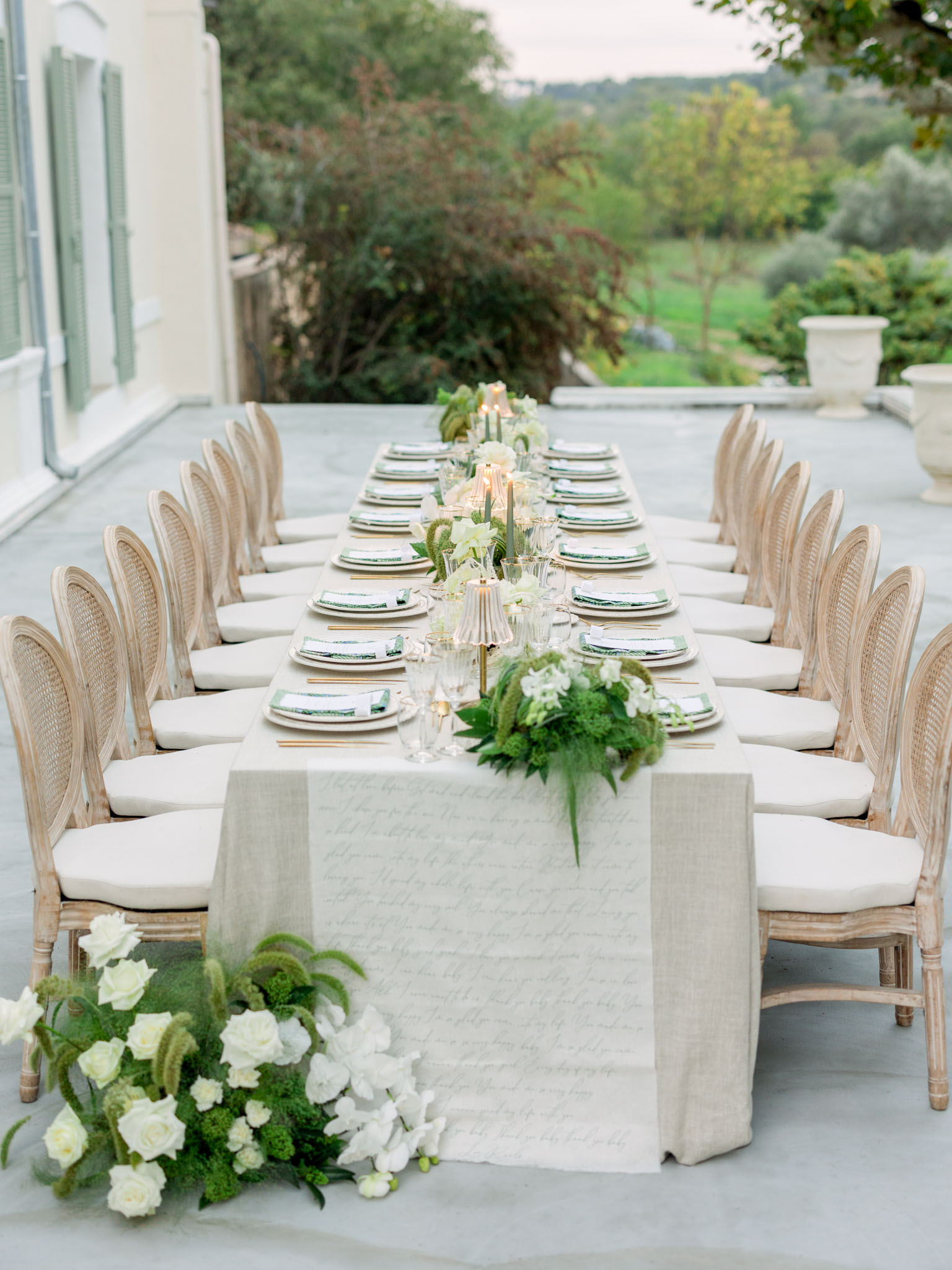 Long reception table on a terrace with white floral centerpieces, rattan chairs, and countryside views