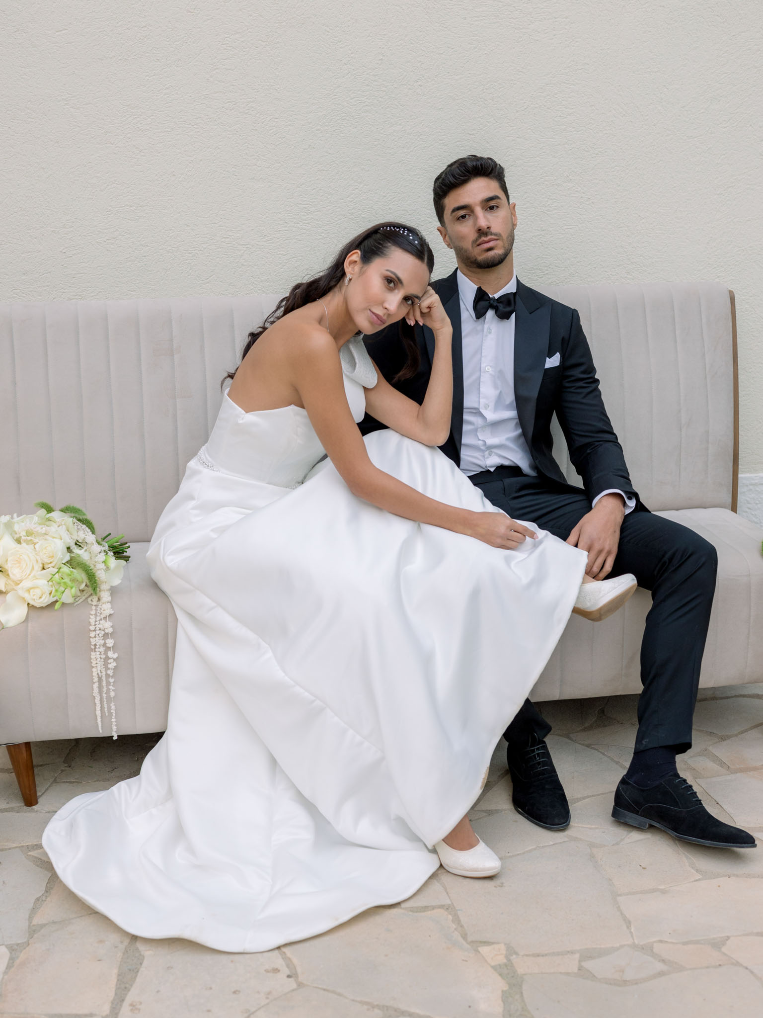 Bride and groom seated on a cream sofa for a formal portrait with bridal bouquet of roses and hydrangea