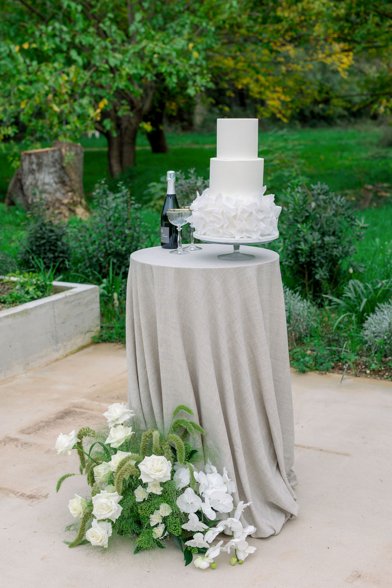Three-tiered white wedding cake on linen-draped table with ivory roses and hydrangeas in outdoor garden setting