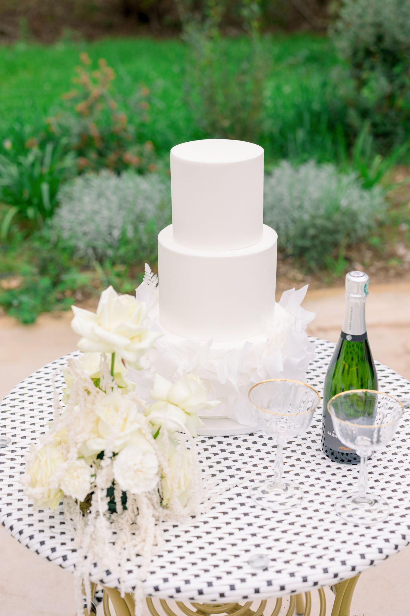 Two-tiered white wedding cake with champagne and ivory rose bouquet on geometric tablecloth at outdoor garden reception