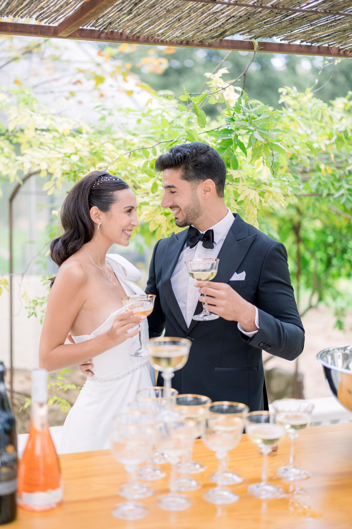 Couple toasting with champagne flutes under vine-draped wooden pergola at outdoor cocktail hour