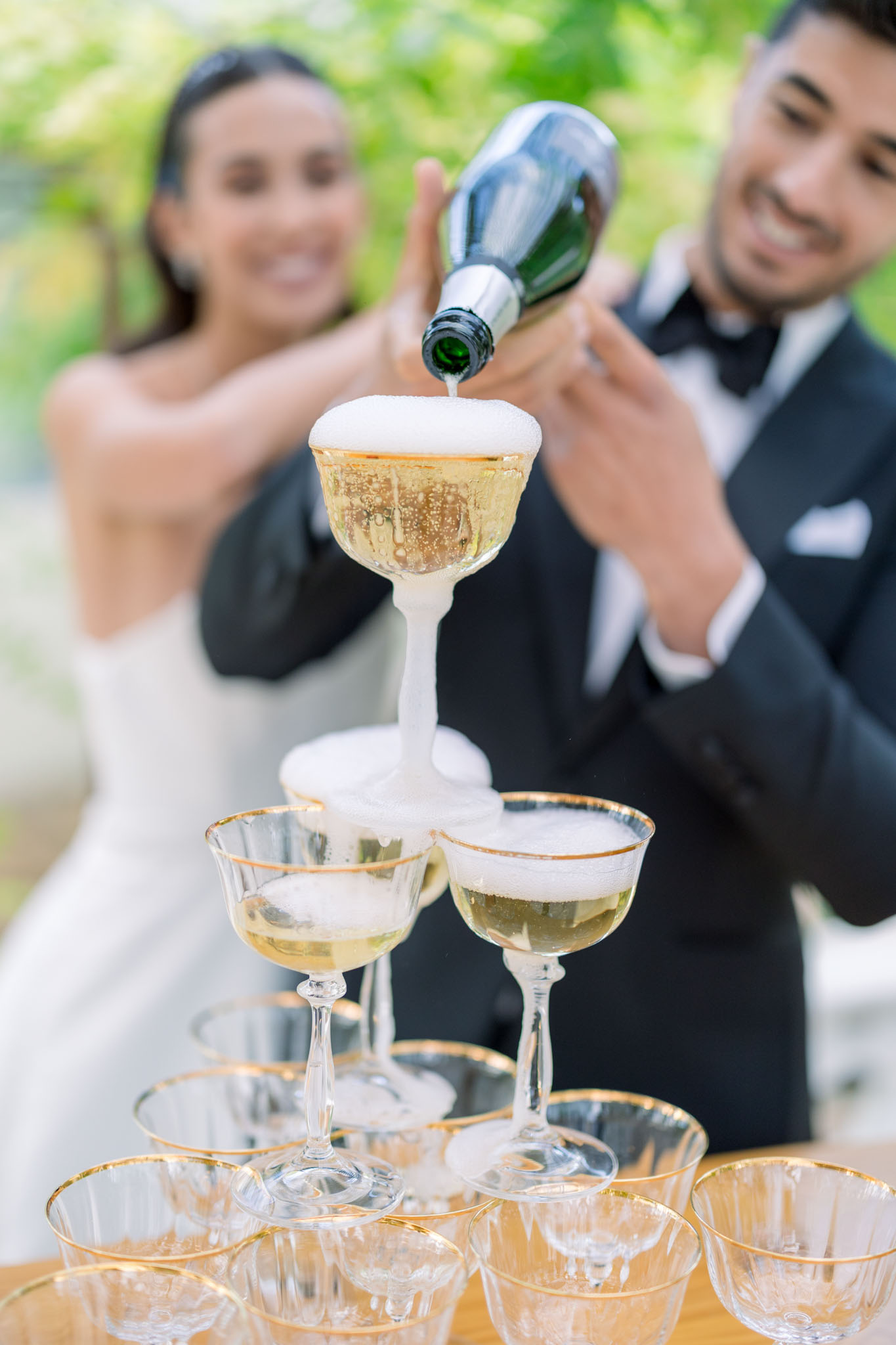 Champagne poured into the top coupe of a pyramid of gold-rimmed glasses; bride and groom blurred in background.