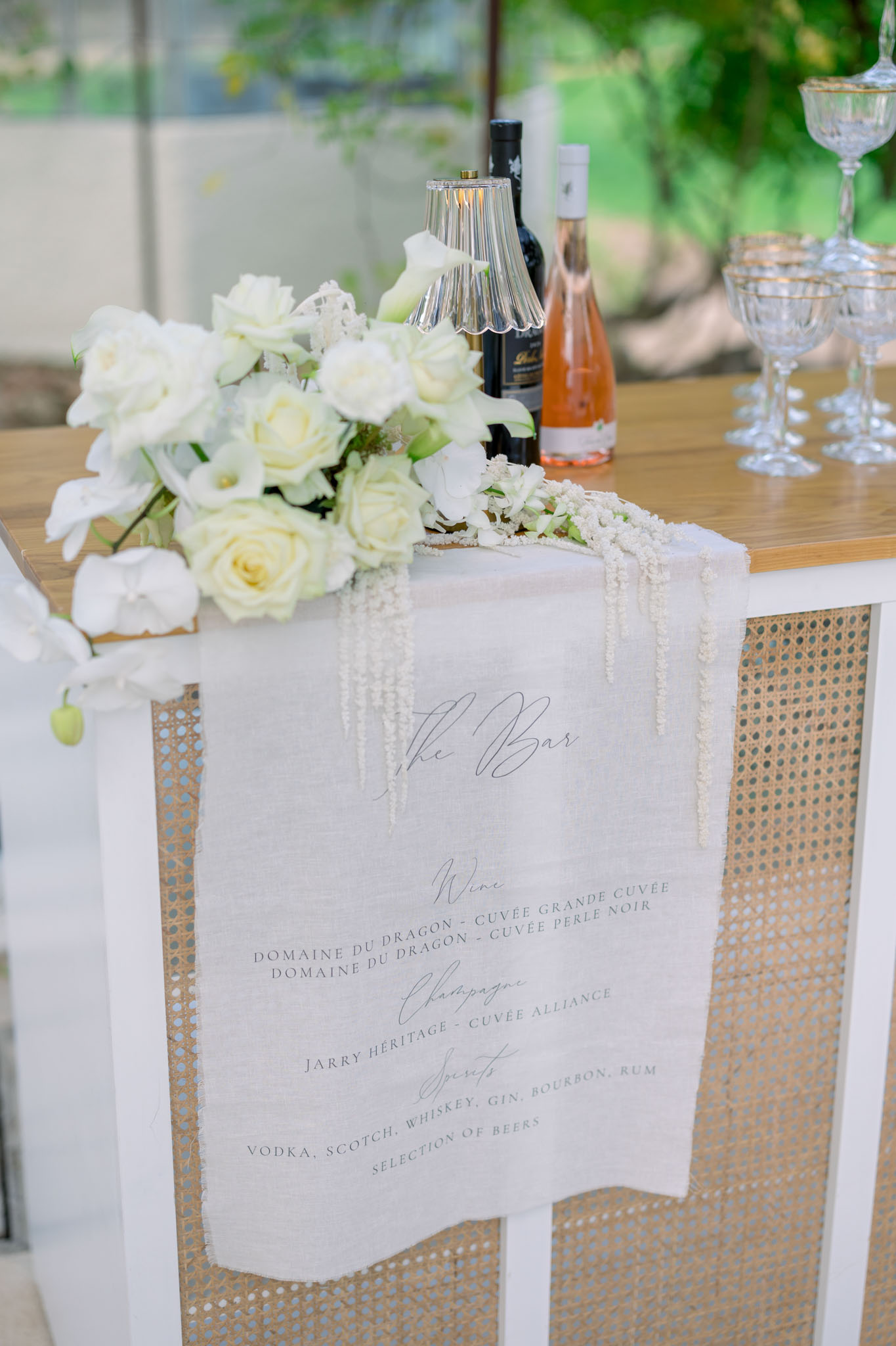 Cocktail bar table with handwritten menu card, ivory and pale yellow roses, wine bottles, and crystal glassware