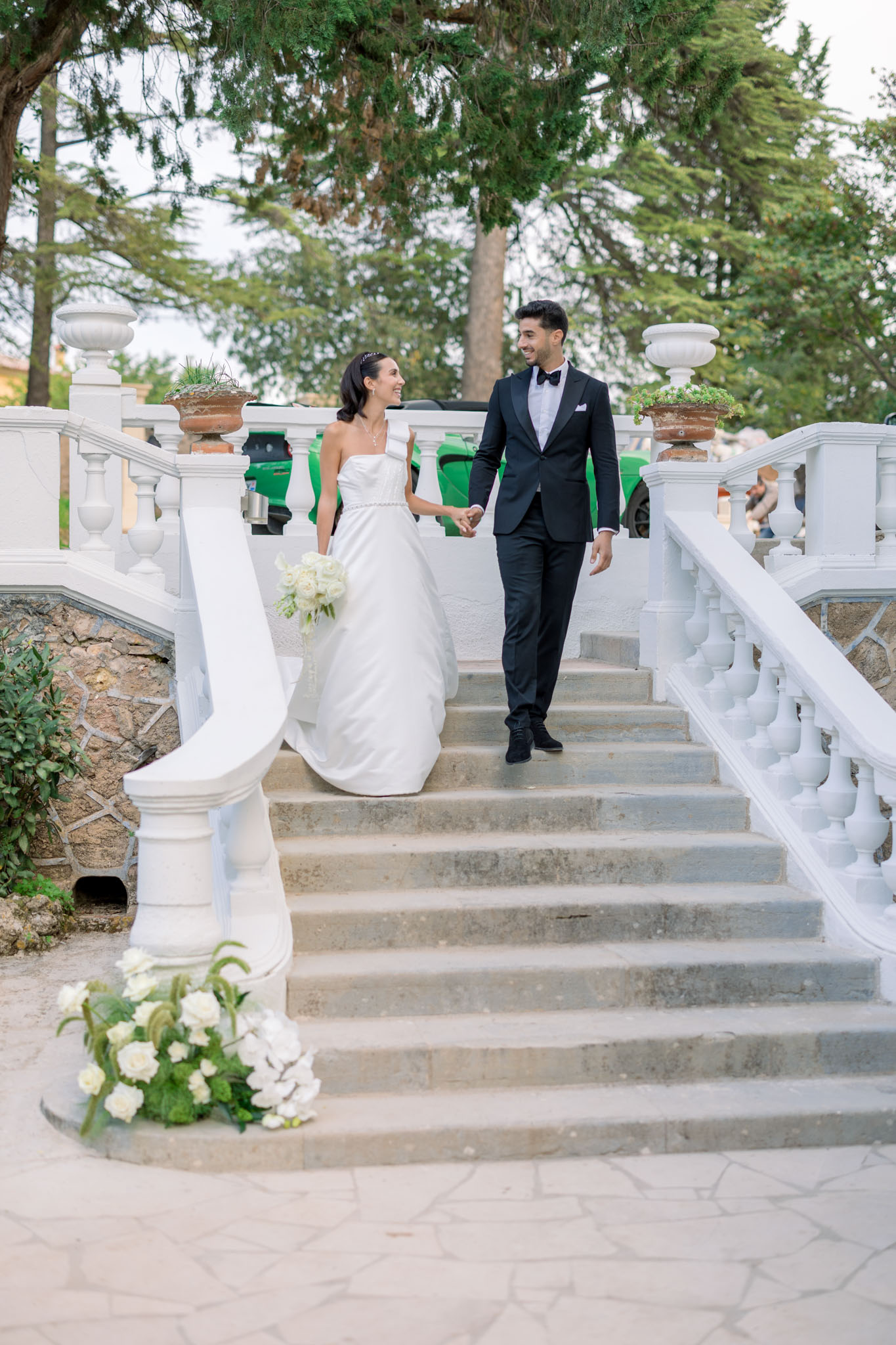 Bride and groom walk hand-in-hand down stone staircase with white balustrades in formal neoclassical garden