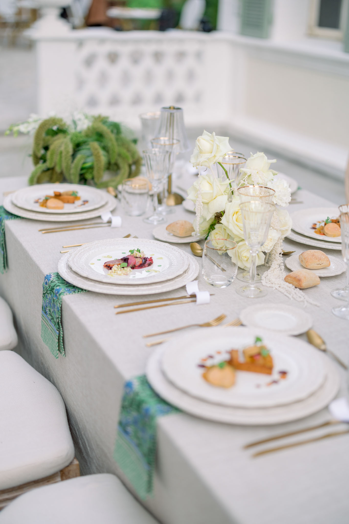 Close-up reception table with plated meal, white roses, emerald napkins, and gold flatware on white linen