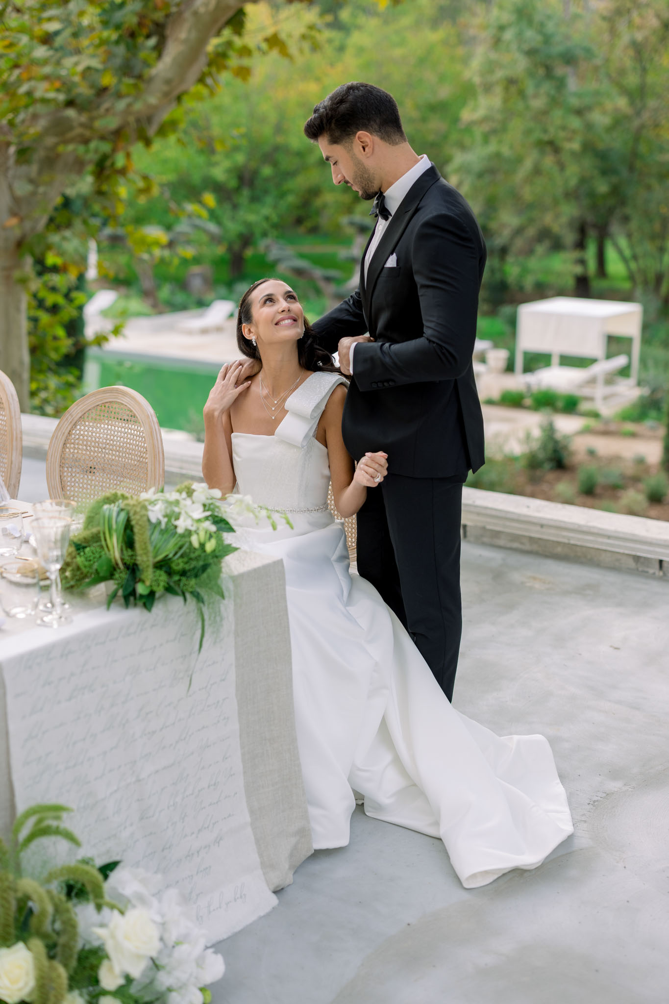 Bride in white column gown and groom in tuxedo at outdoor reception table with white floral centerpiece