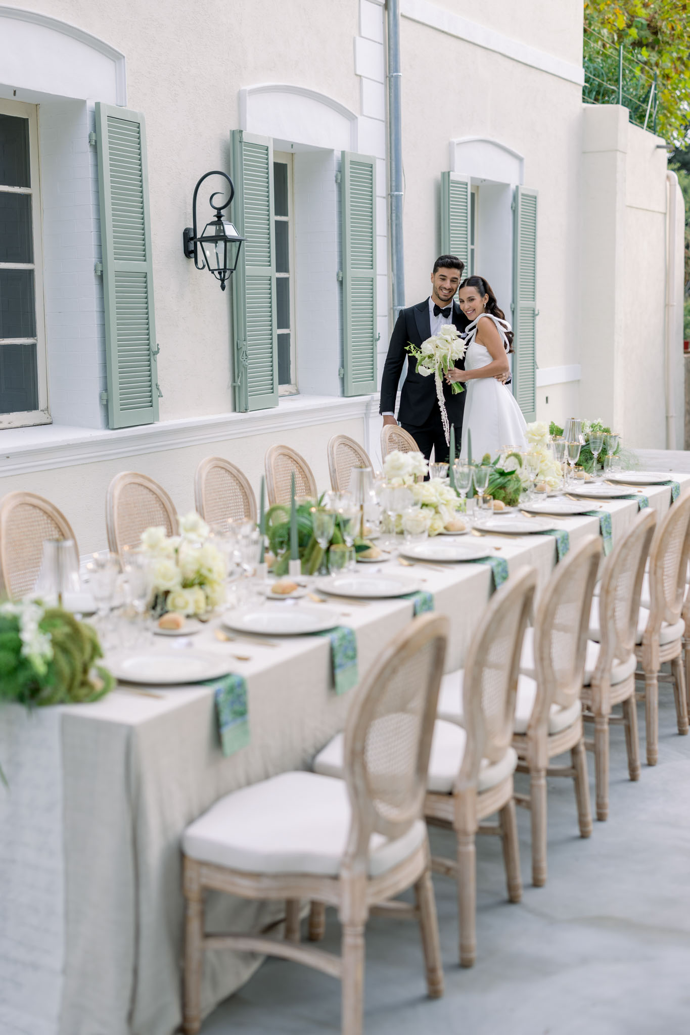 Outdoor reception table with ivory linen and sage green napkins, couple in background beside Mediterranean building