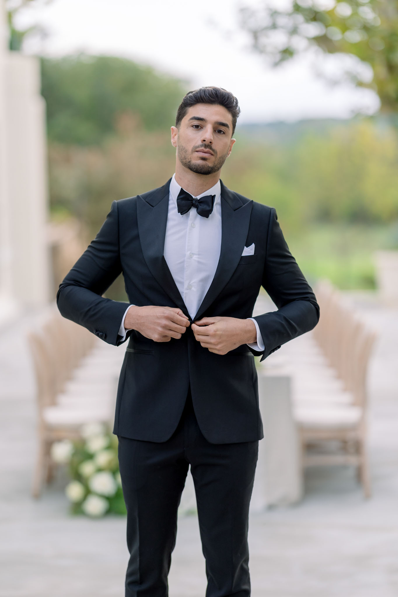 Groom in black tuxedo adjusting jacket on stone pathway beside a château building with soft-focus greenery behind