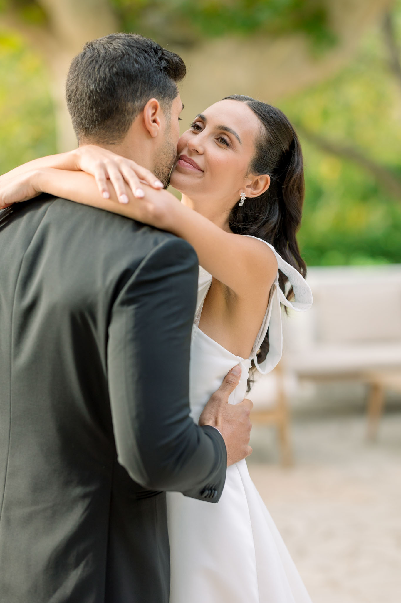 Bride and groom embracing outdoors; bride in white V-neck long-sleeve gown, groom in charcoal suit, green foliage behind.