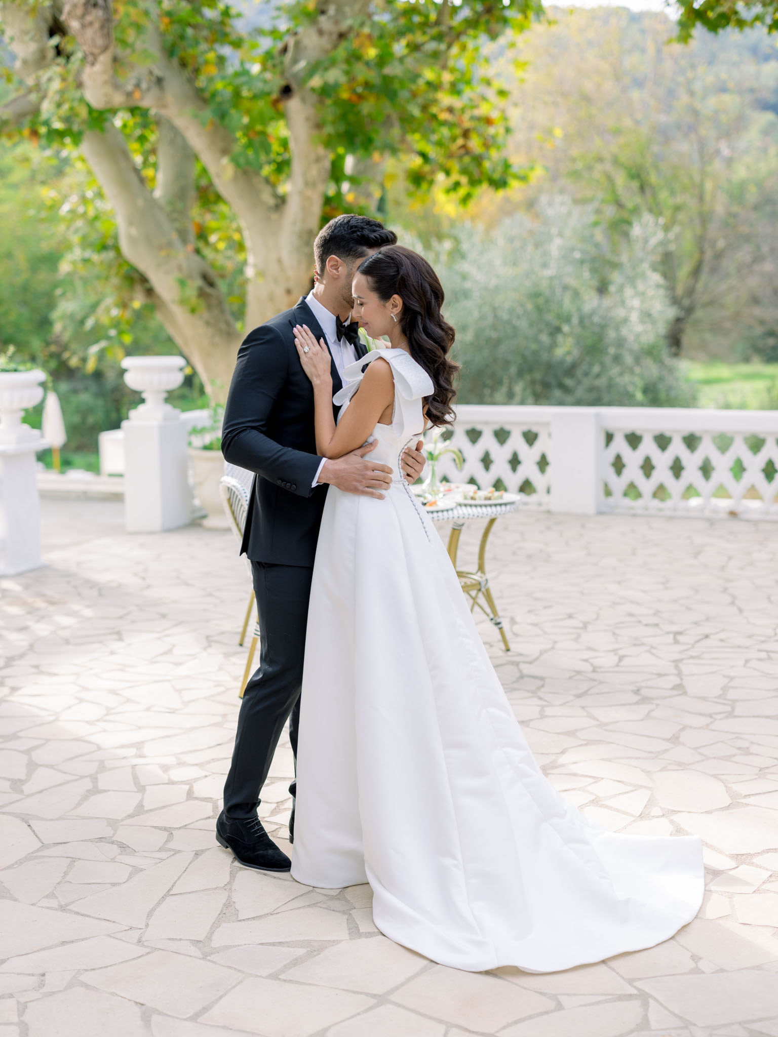 Groom kissing bride's temple on stone balustrade terrace with Versailles planters and gold bistro chairs