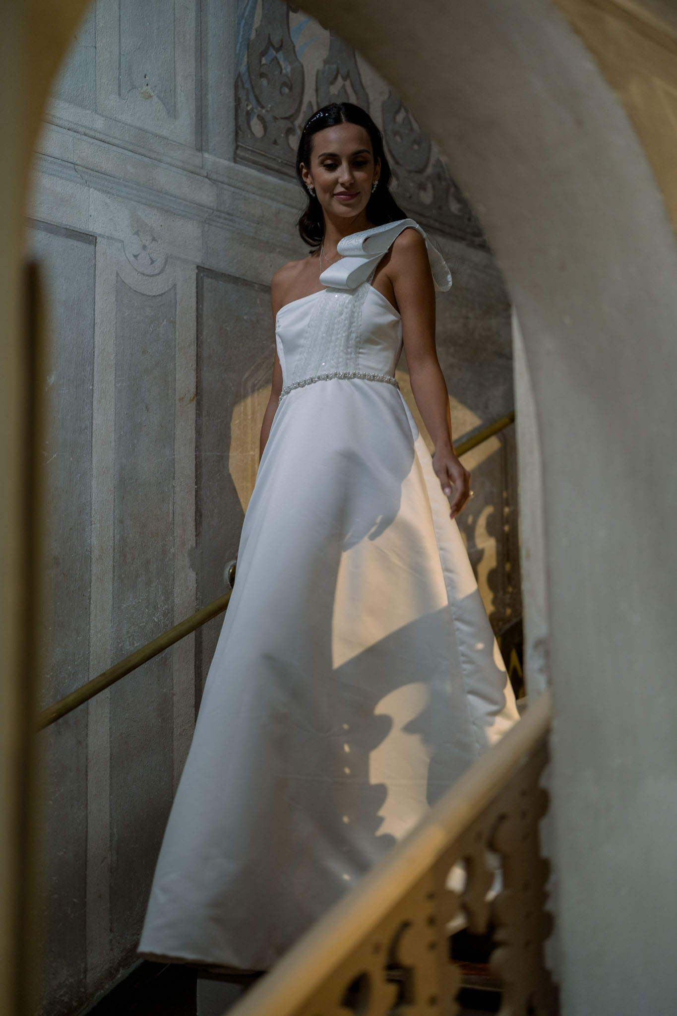 Bride in white one-shoulder gown in narrow stone corridor with dramatic light casting shadow on carved walls