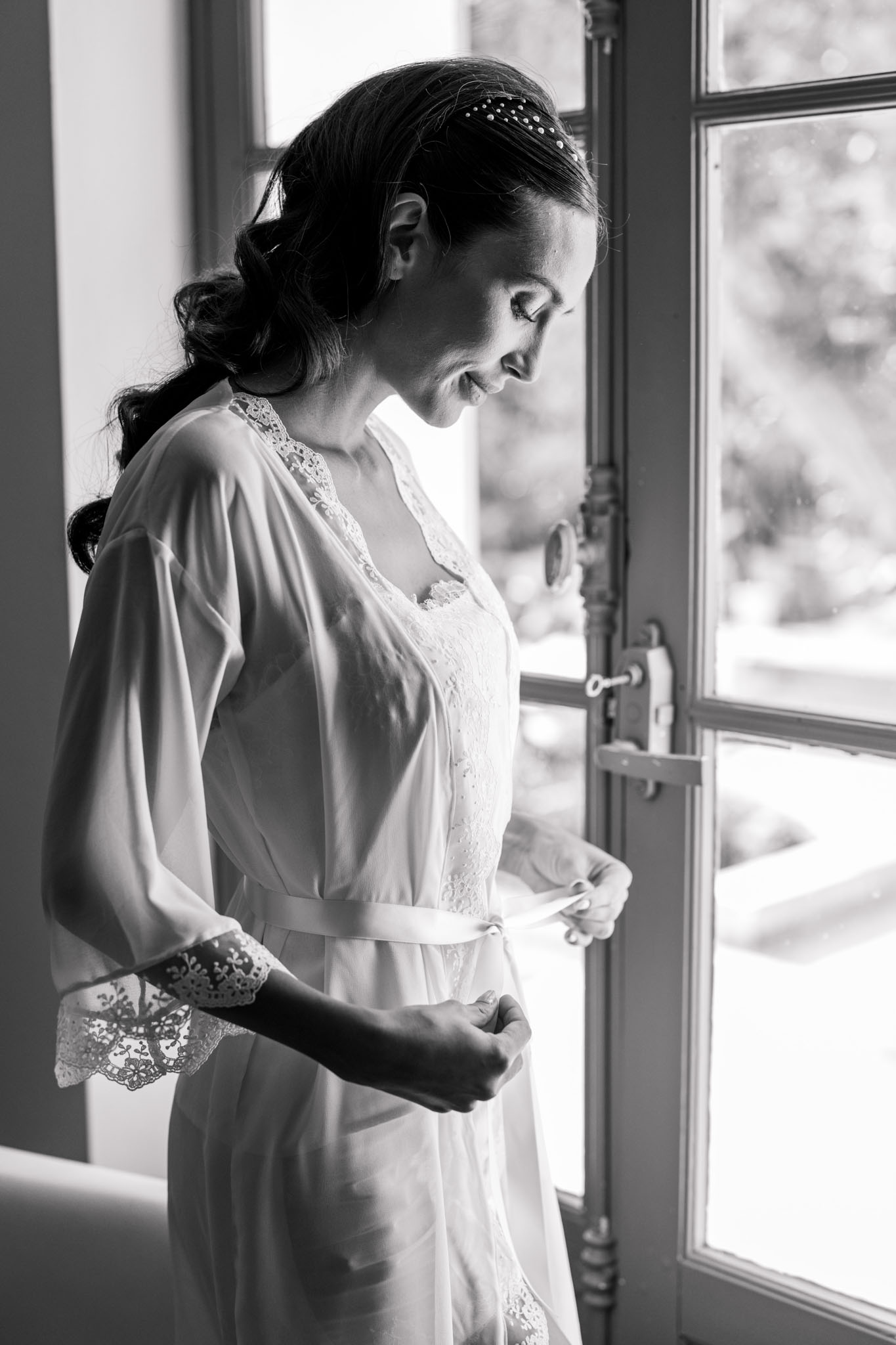Bride in white lace-trim robe at window with pearl-embellished low bun, looking downward, black and white photo