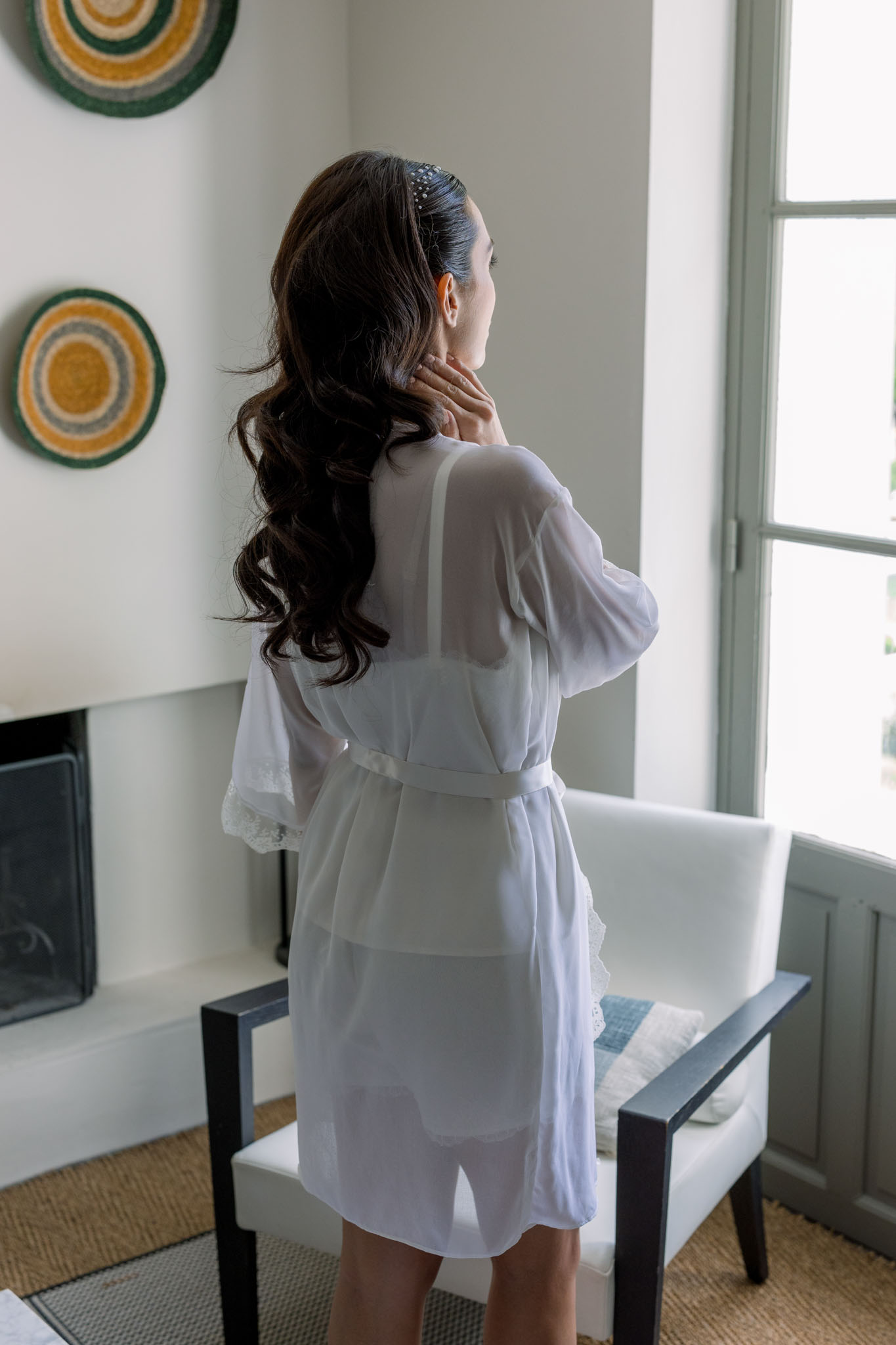 Bride in white silk robe gazing out window with beaded hair accessory during getting ready