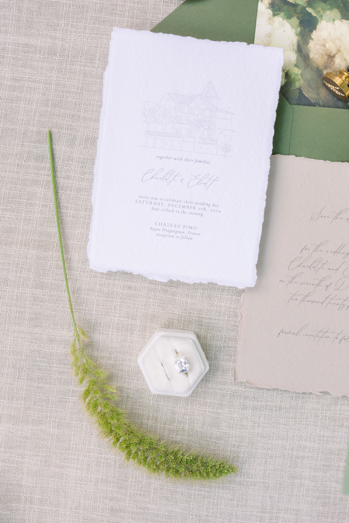 Flat lay of handmade paper wedding invitation with fern, white astilbe, and diamond engagement ring on linen