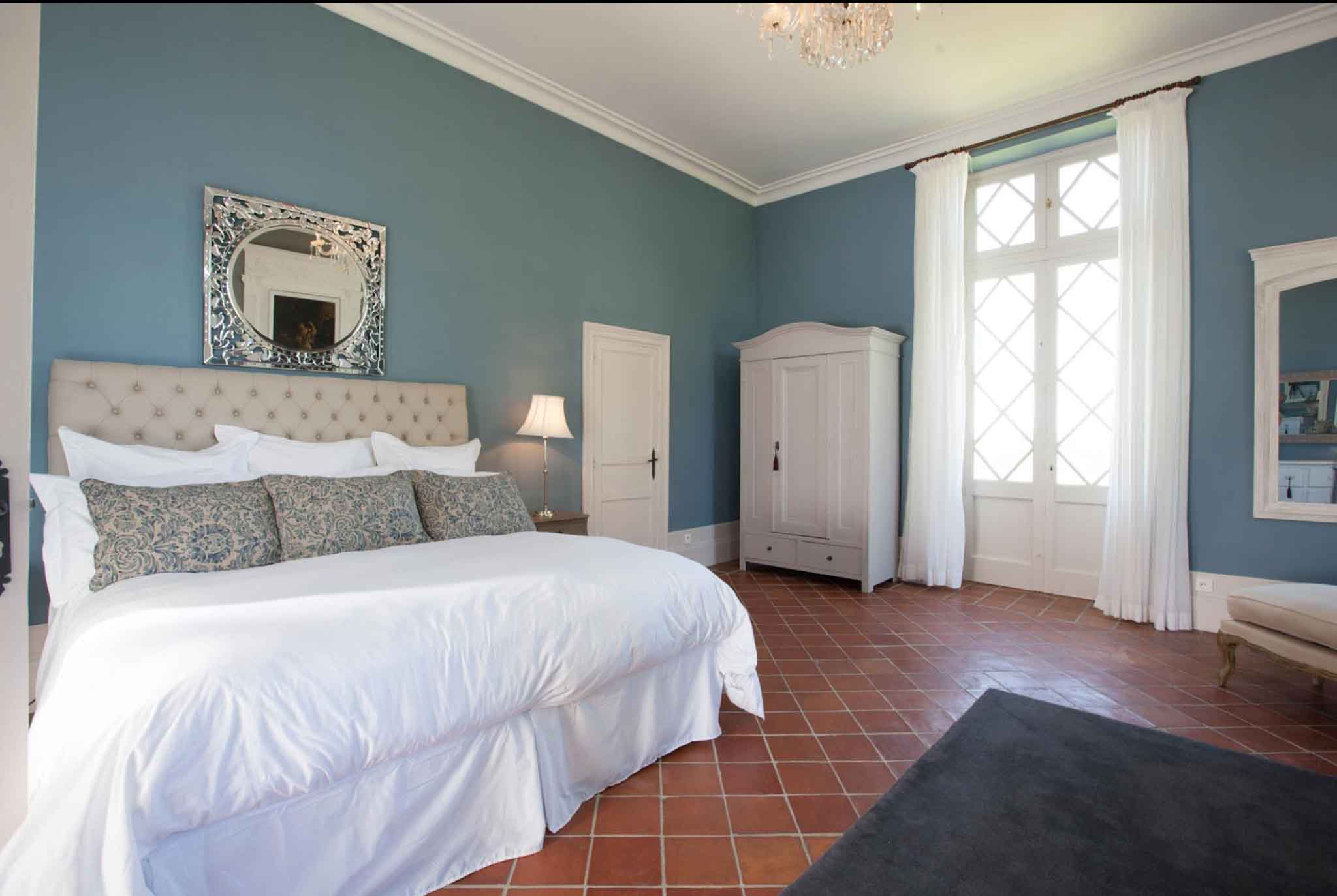 Chateau bedroom with steel-blue walls, crystal chandelier, tufted headboard, and white shuttered windows