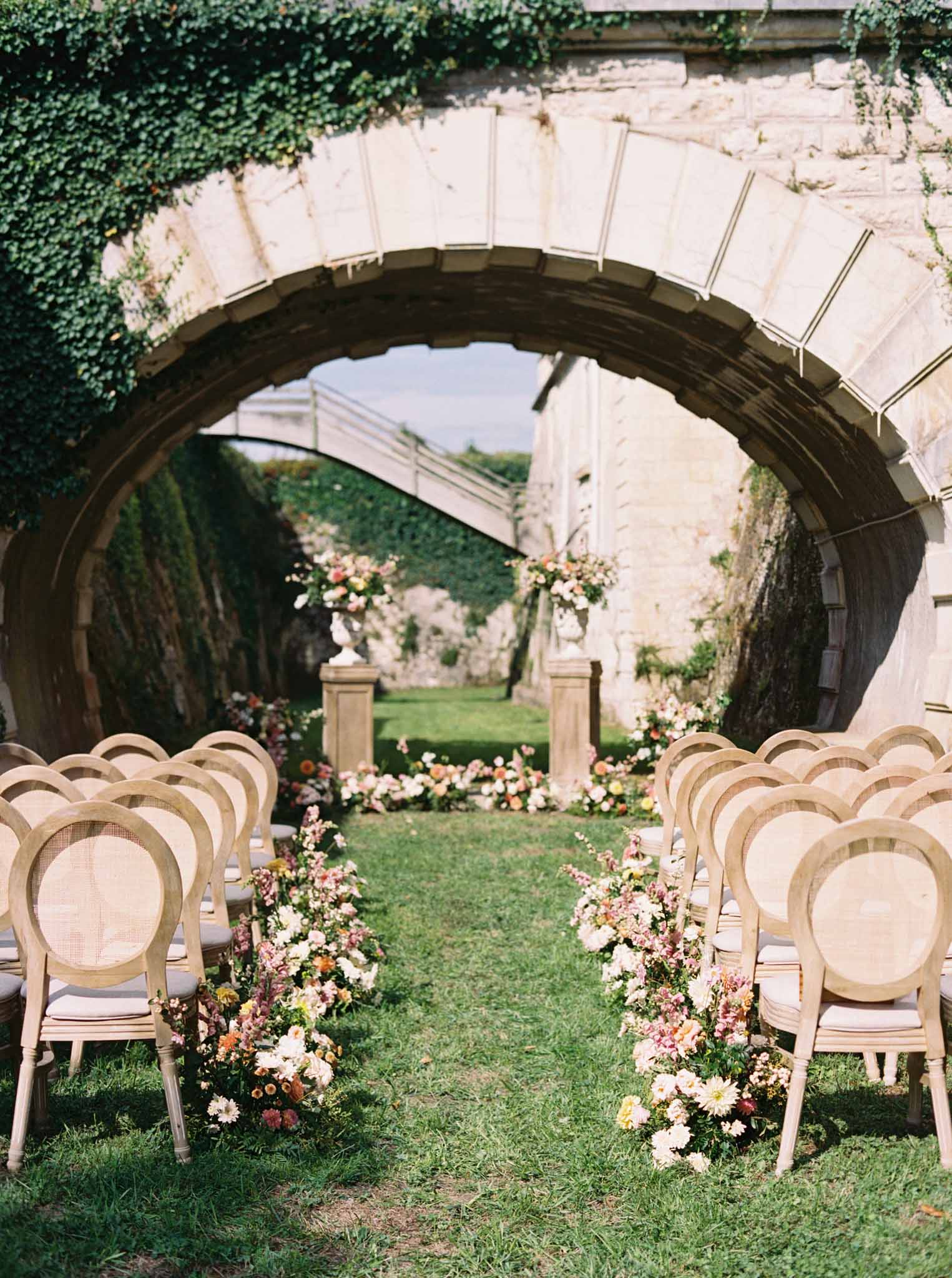 Outdoor ceremony aisle under ivy-draped stone arch with cream and coral floral arrangements on pedestals