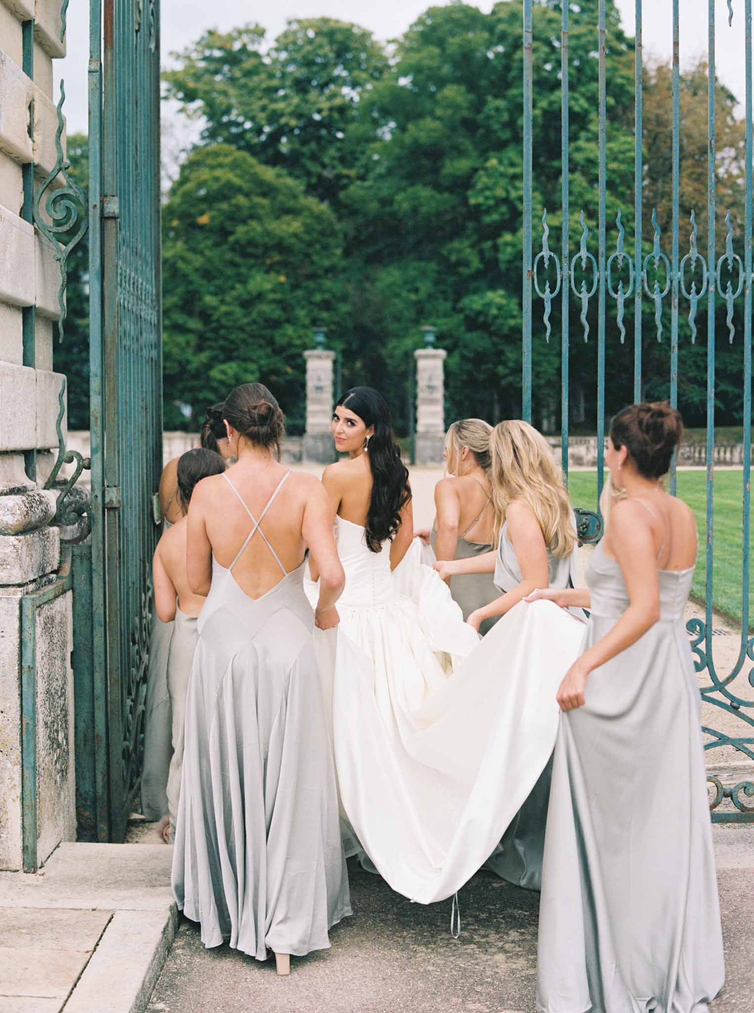 Bride and five bridesmaids in neutral gowns photographed from behind at ornate teal iron gates