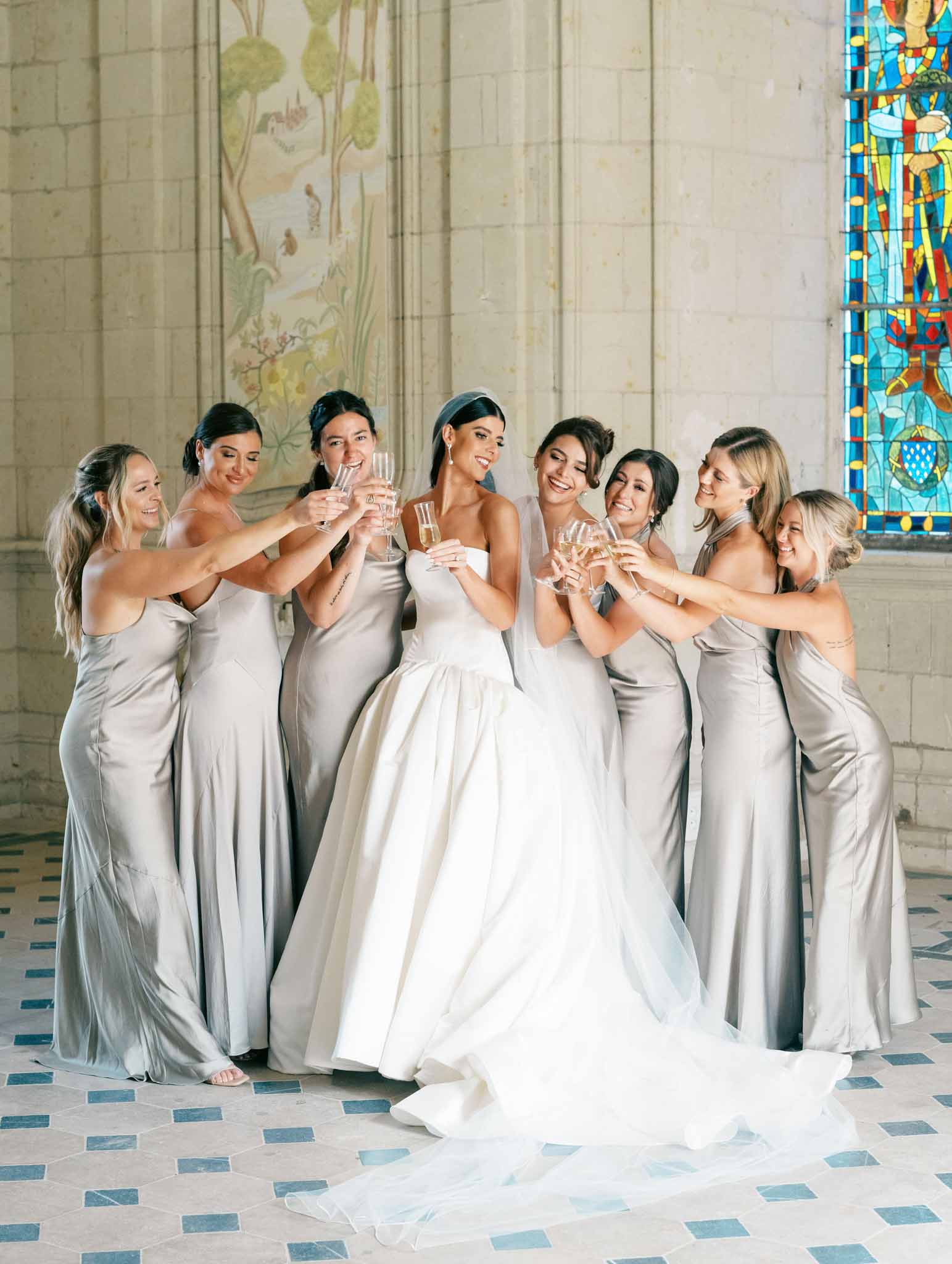 Bride in ivory ball gown with eight bridesmaids in taupe slip dresses toasting in chapel with blue stained glass