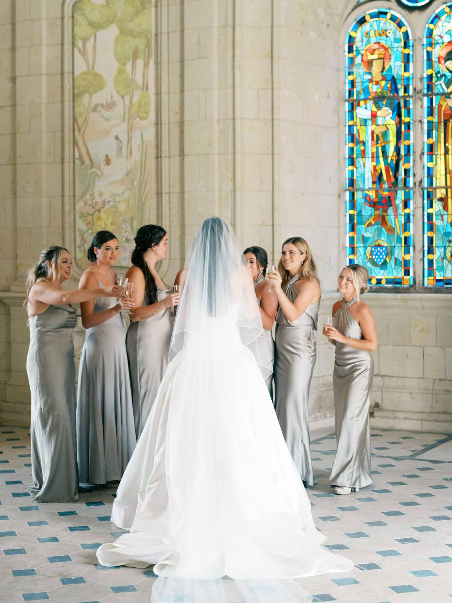 Bride from behind in chapel aisle with six bridesmaids in sage green gowns raising champagne flutes