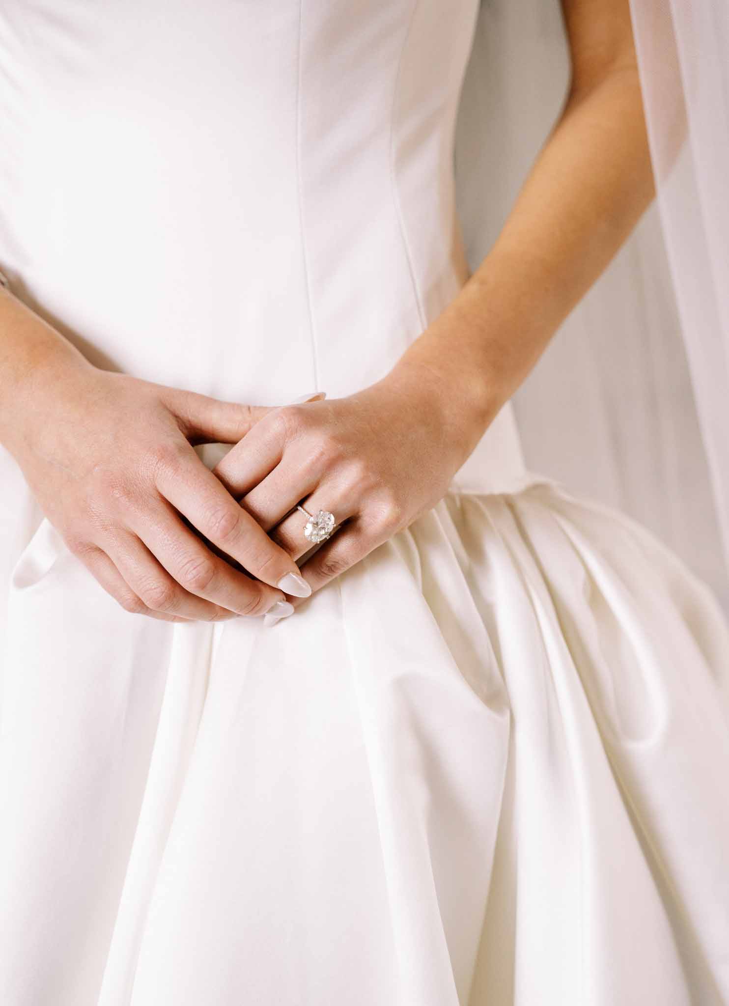 Close-up of bride's hands resting on ivory silk pleated skirt showing diamond solitaire engagement ring and wedding band