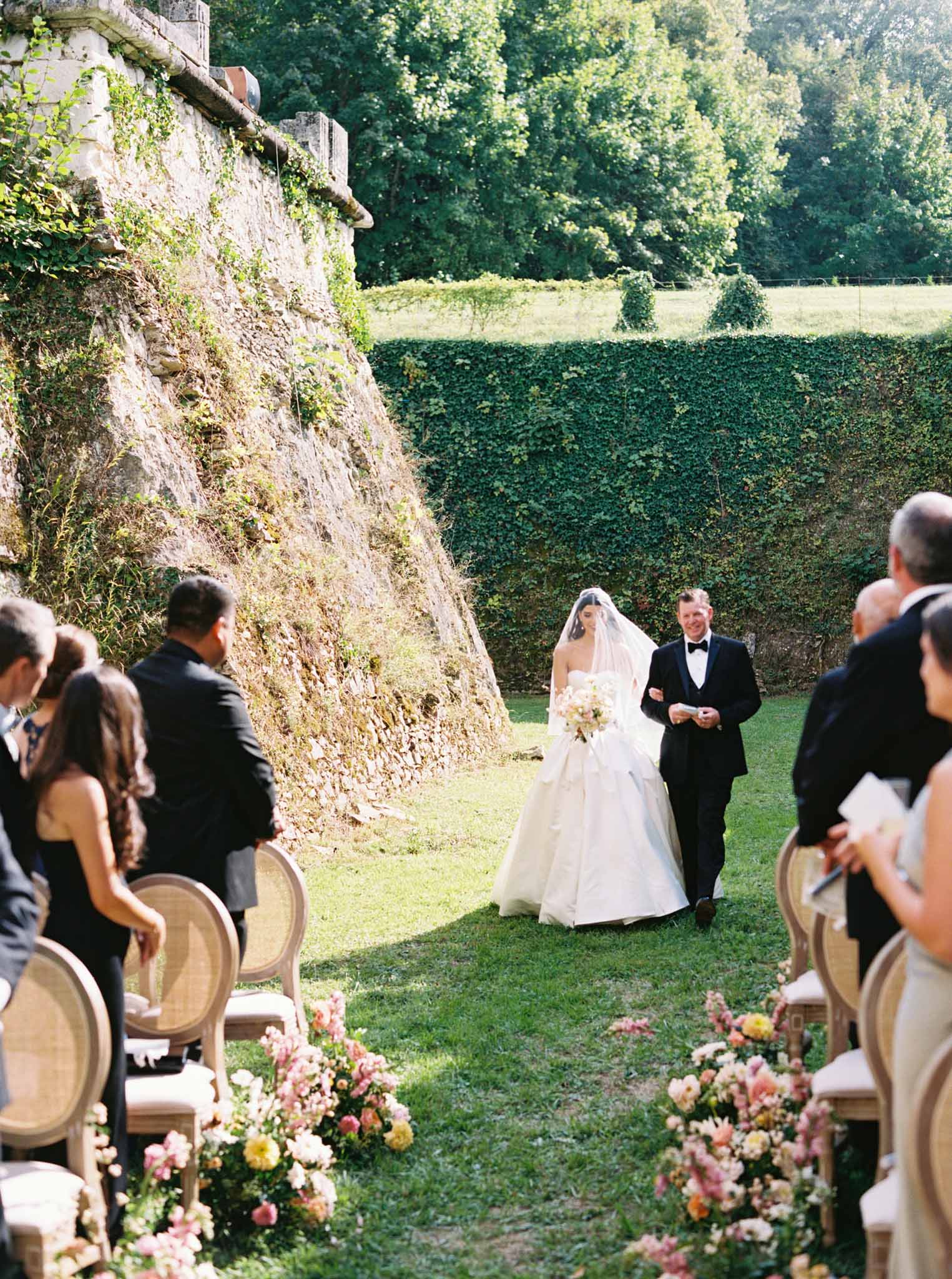 Bride in white ballgown walking aisle arm-in-arm with father past coral and blush floral arrangements at stone château