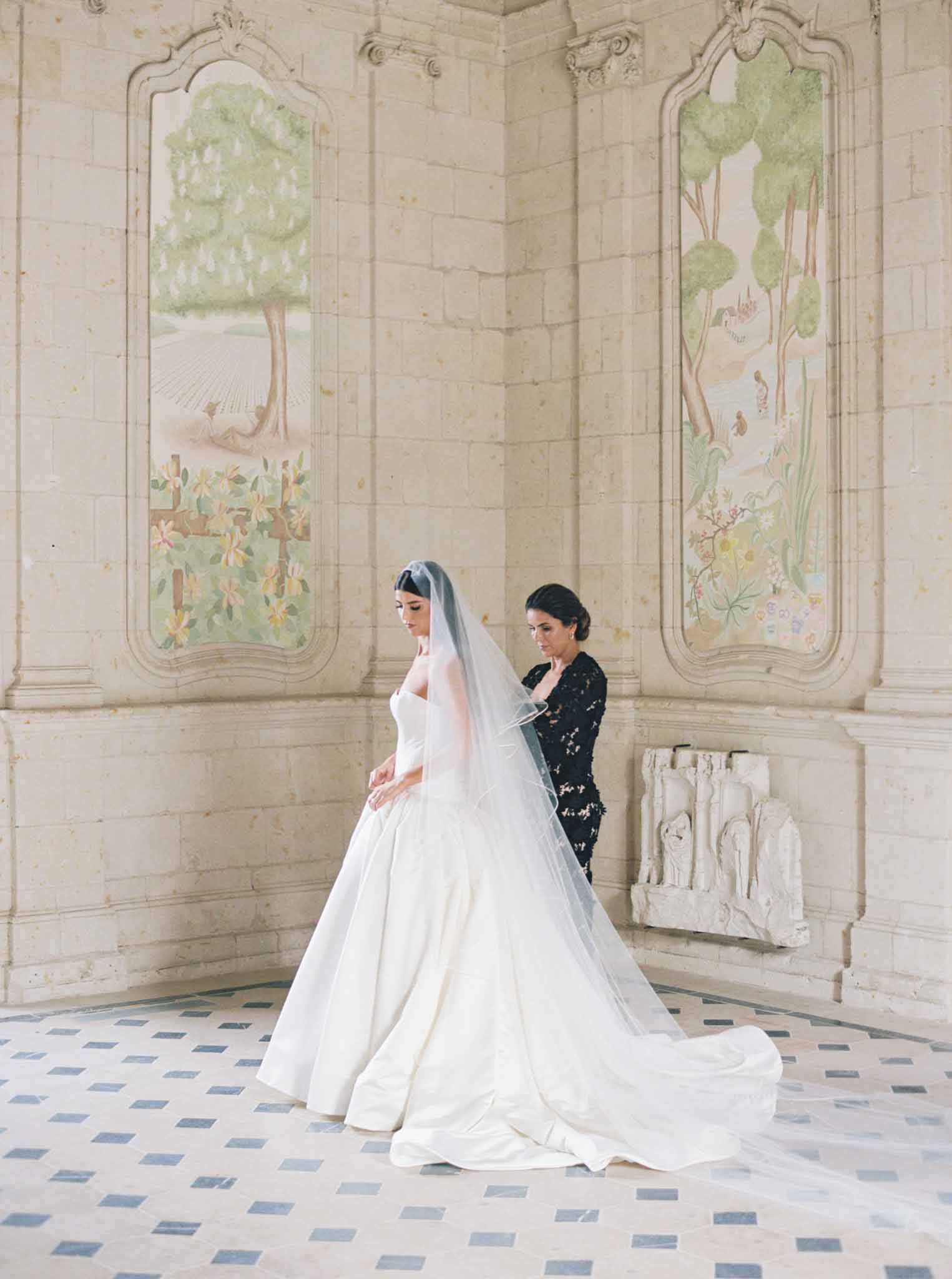 Bride in ivory long-sleeve wedding dress being attended to in ornate room with botanical panel murals and geometric floor