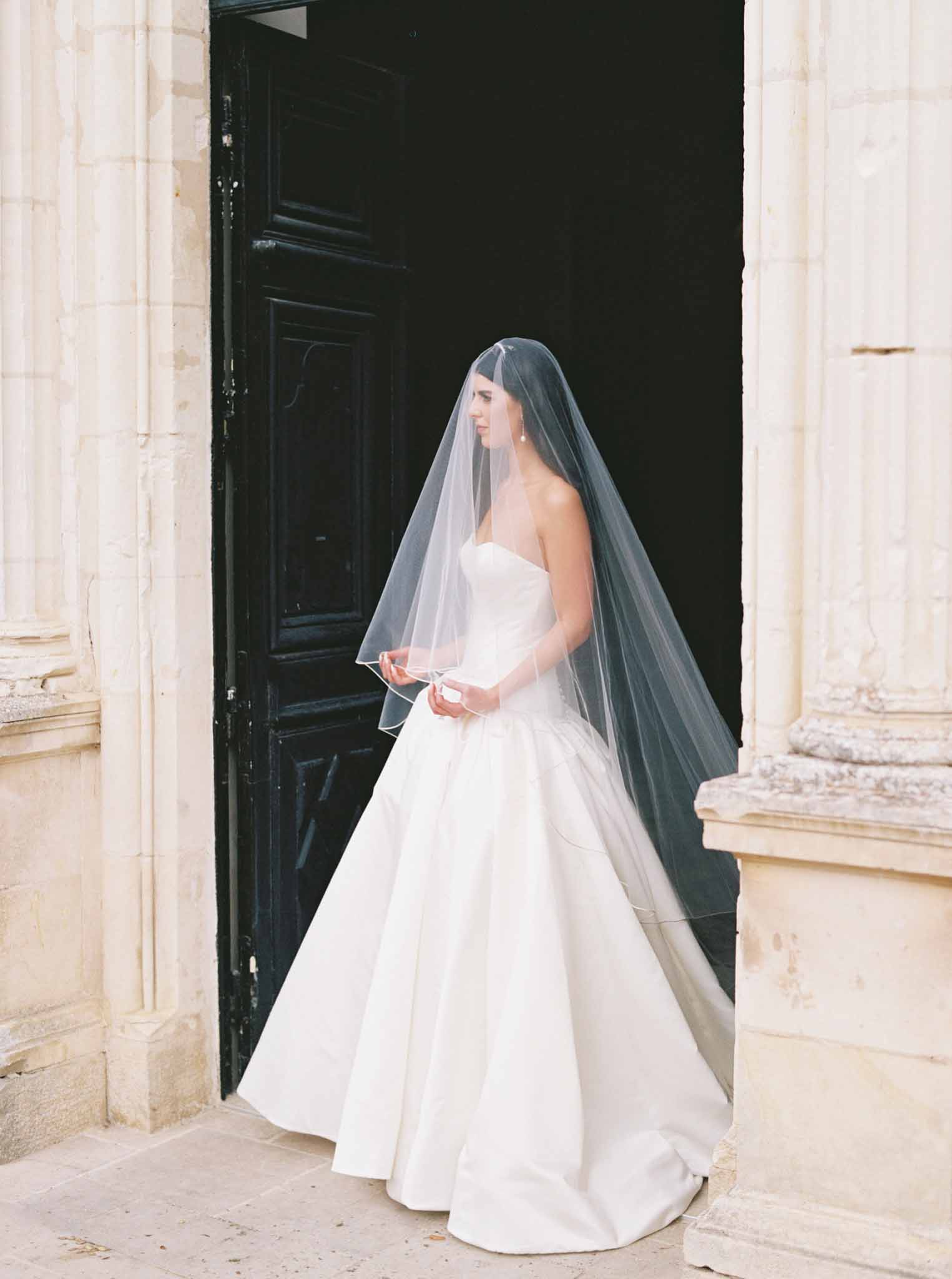 Bride in strapless ball gown and cathedral veil standing before classical stone archway entrance