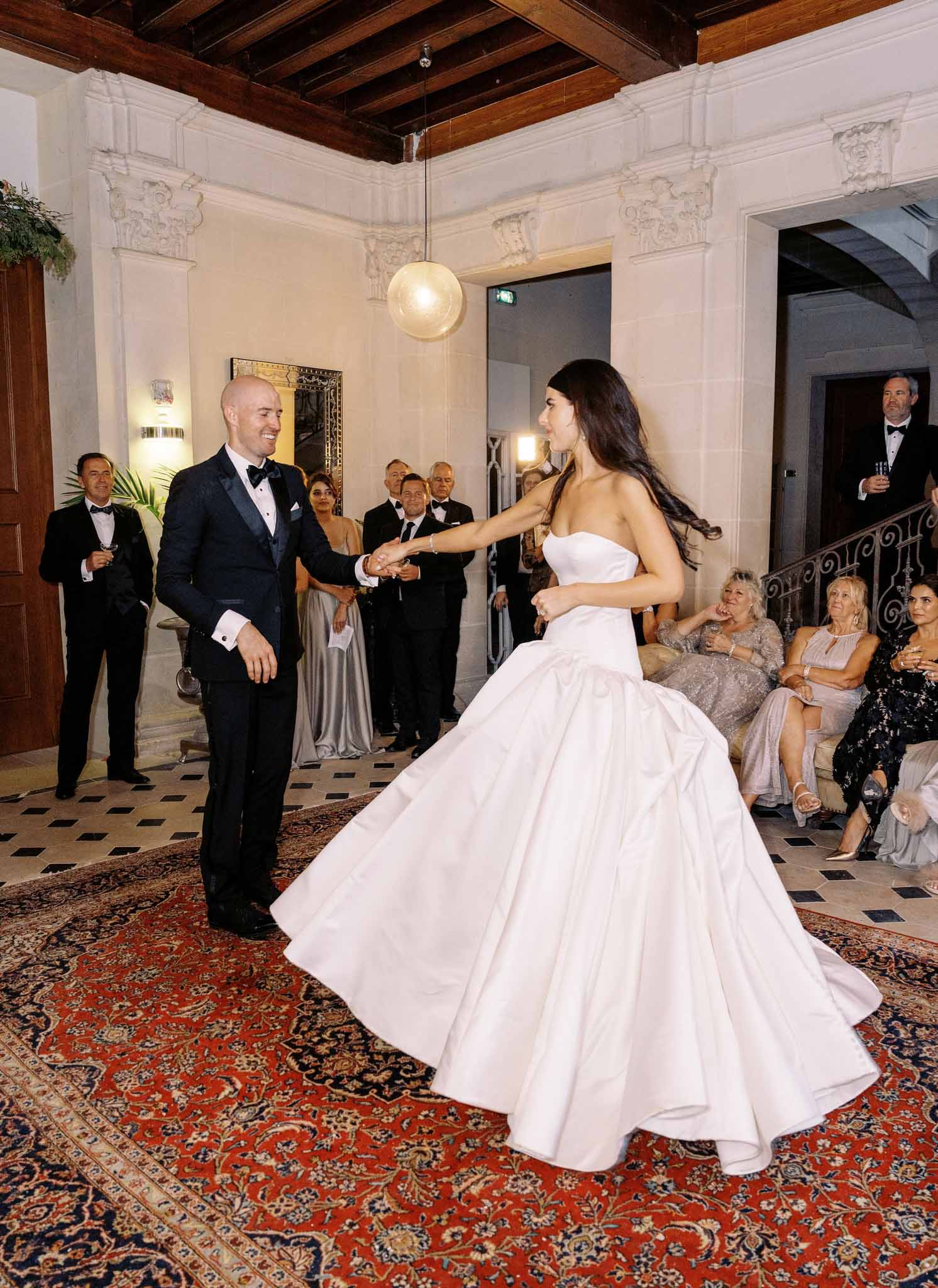 Couple sharing first dance in classical ballroom with ornate corbels, dark beams, and Oriental rug, guests watching from sides