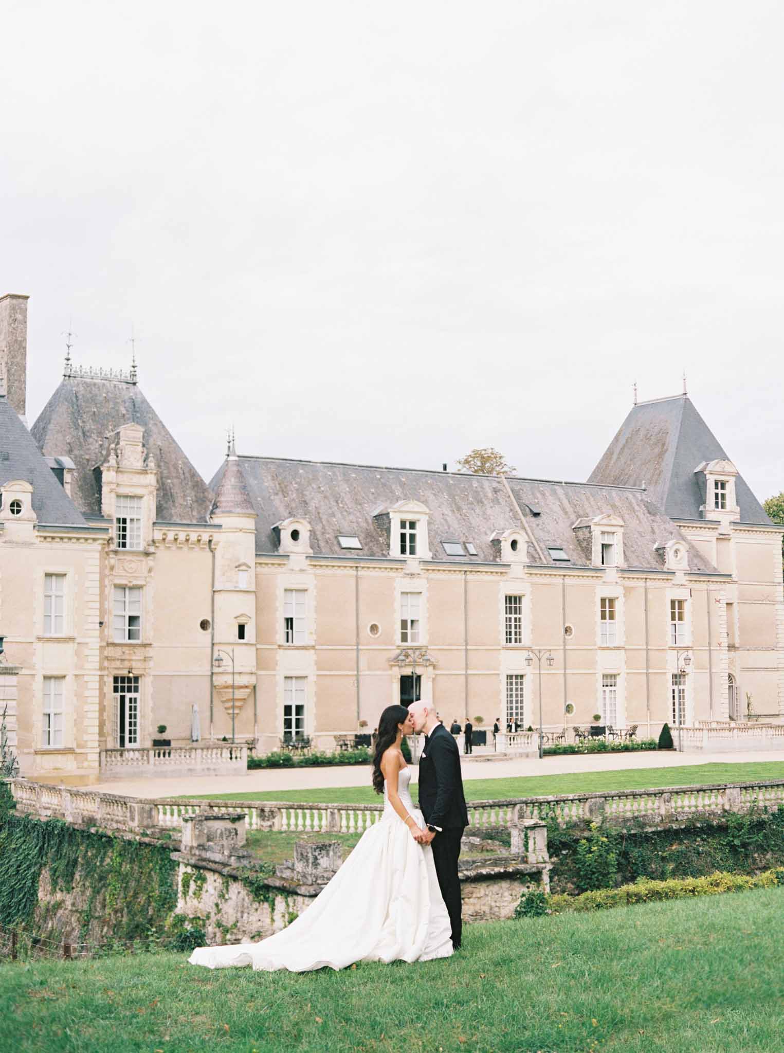Bride and groom kissing on the lawn in front of a grand French château with cream-stone façade and mansard roofline.