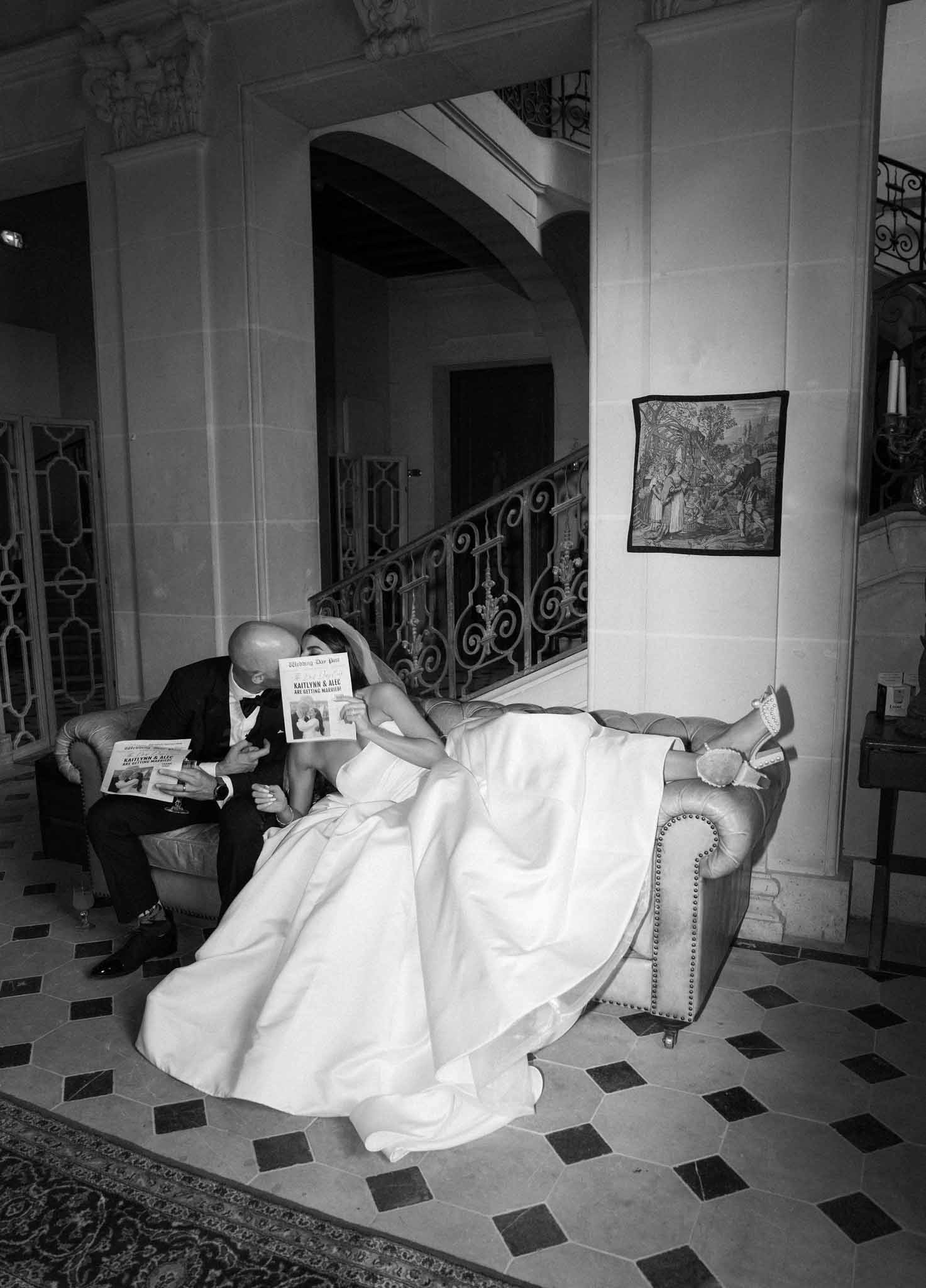 Black and white photo of bride and groom holding newspapers on wingback chair in neoclassical interior