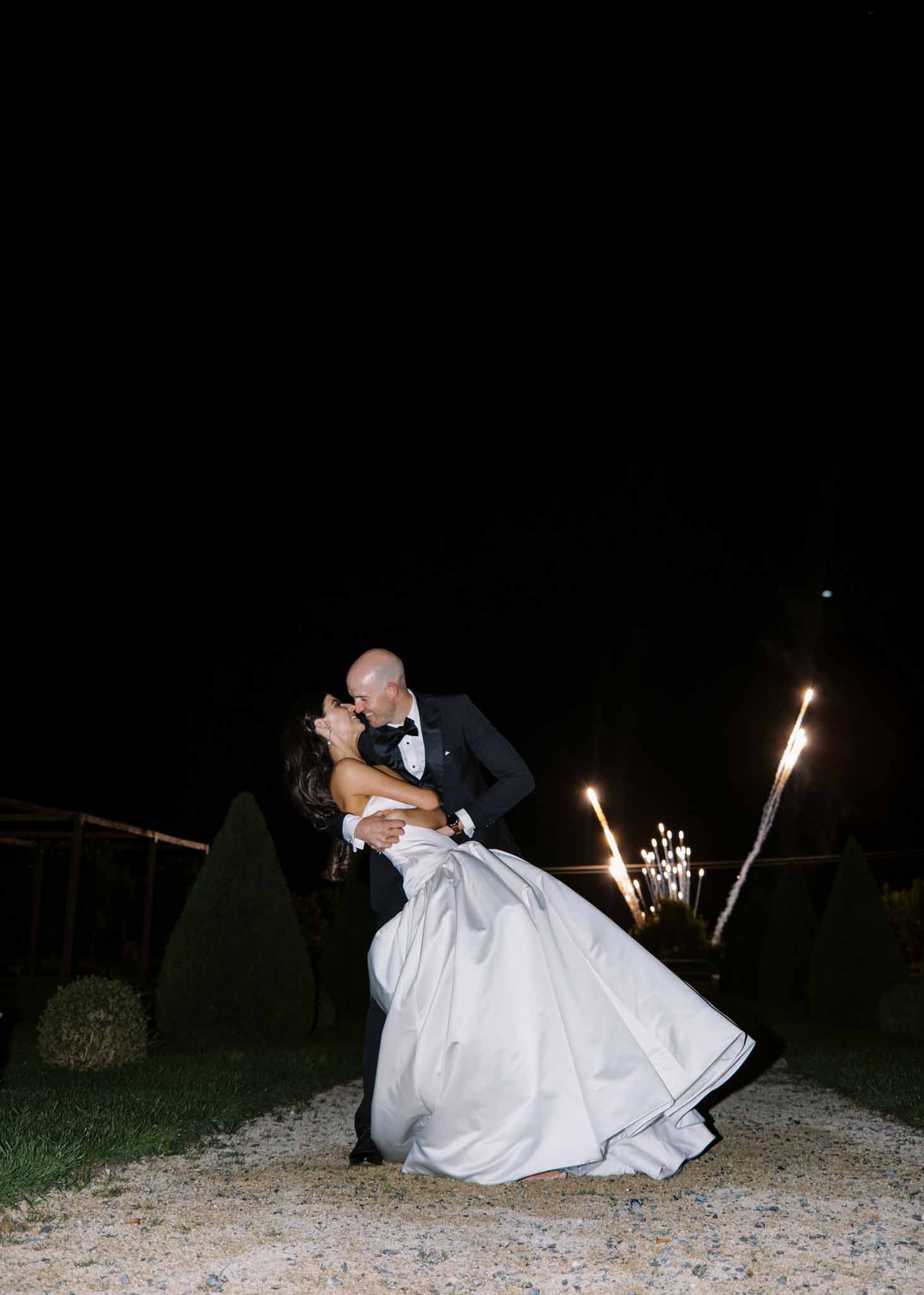 Groom dips and kisses bride in ivory ball gown on gravel path with lit torches and dark night sky behind