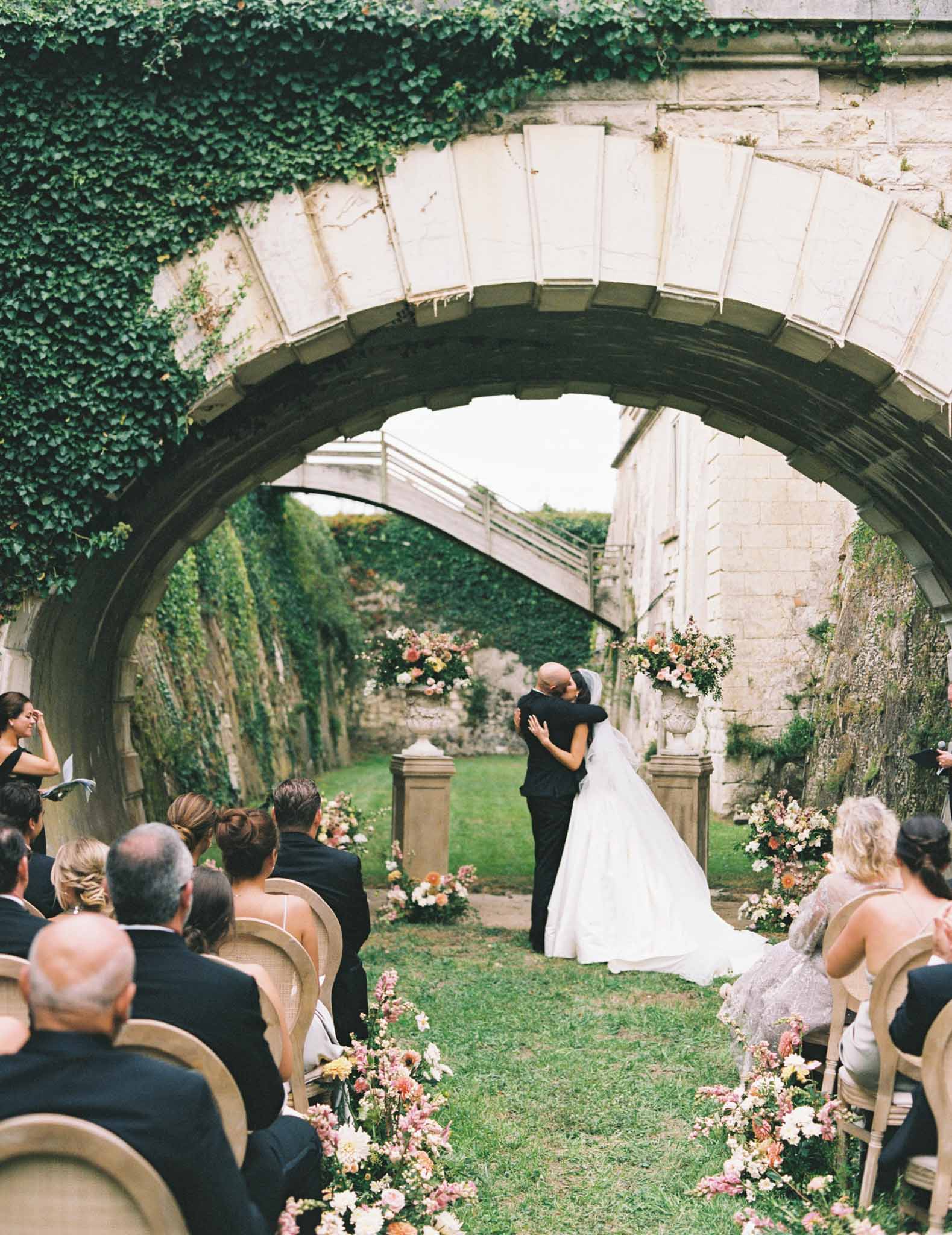 Bride and groom embrace under ivy-covered stone archway in walled garden ceremony, peach florals lining the aisle