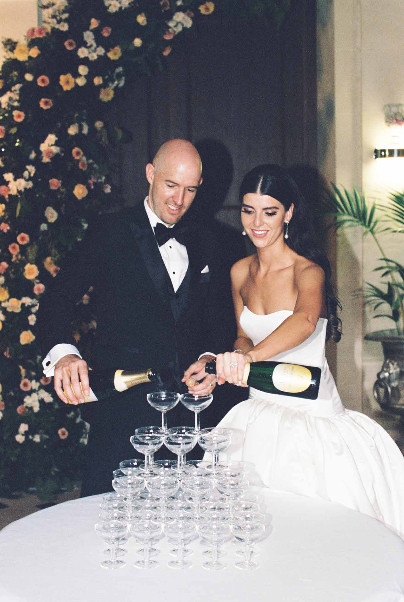 Bride and groom pour champagne into a coupe glass tower in front of a navy floral backdrop at their reception
