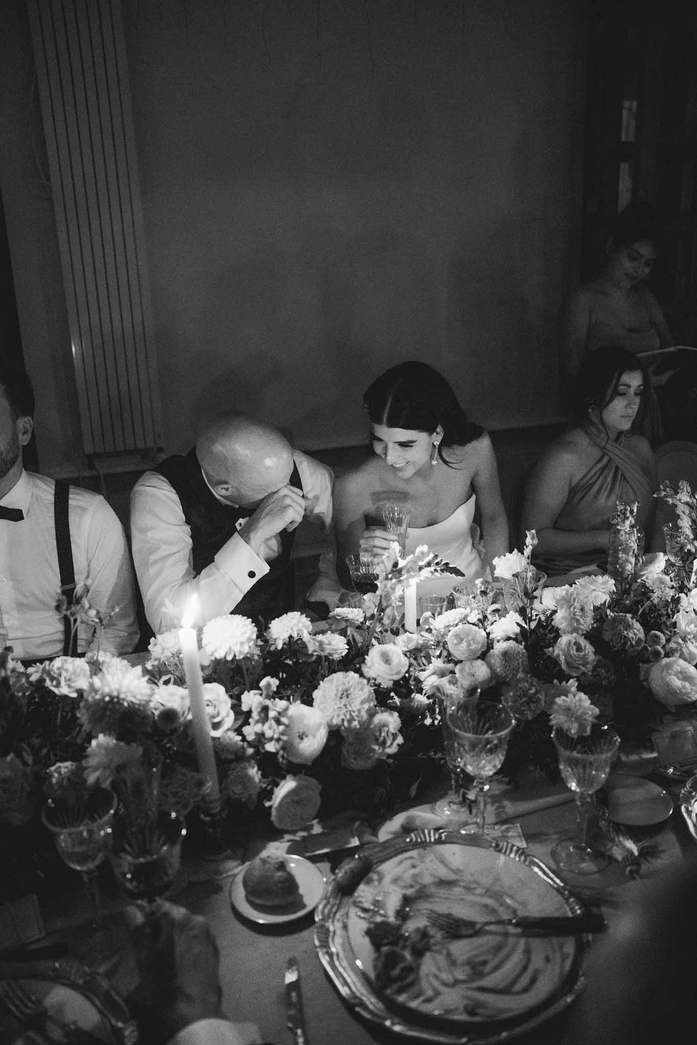 Groom leaning toward bride in a tender moment at the reception dinner table; floral centerpiece of roses and dahlias in foreground.