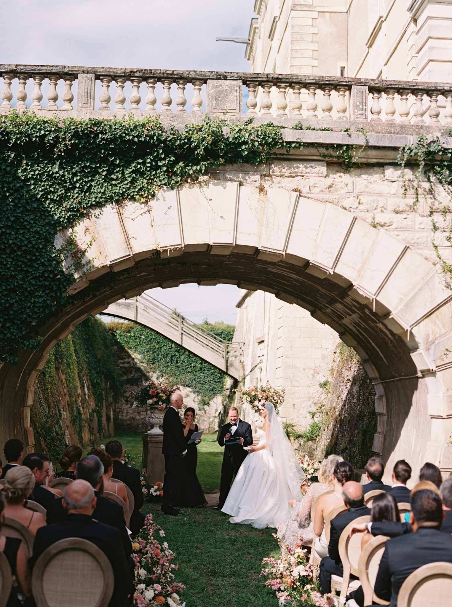 Wedding ceremony under stone arch in ivy courtyard, 50-60 guests on wood chairs, white fabric panel backdrop above couple