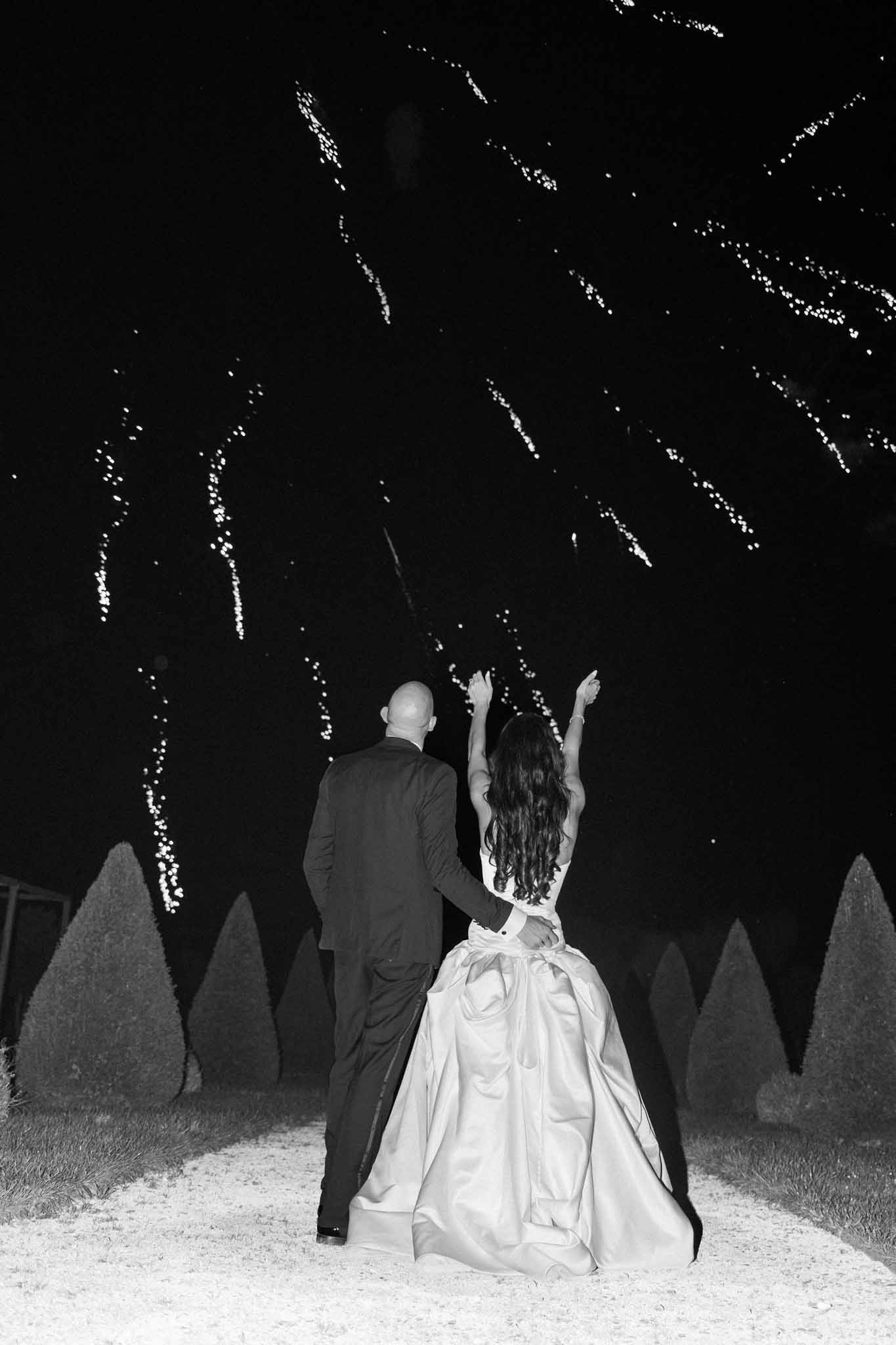 Black and white photo of bride and groom on garden path at night with string lights and topiary