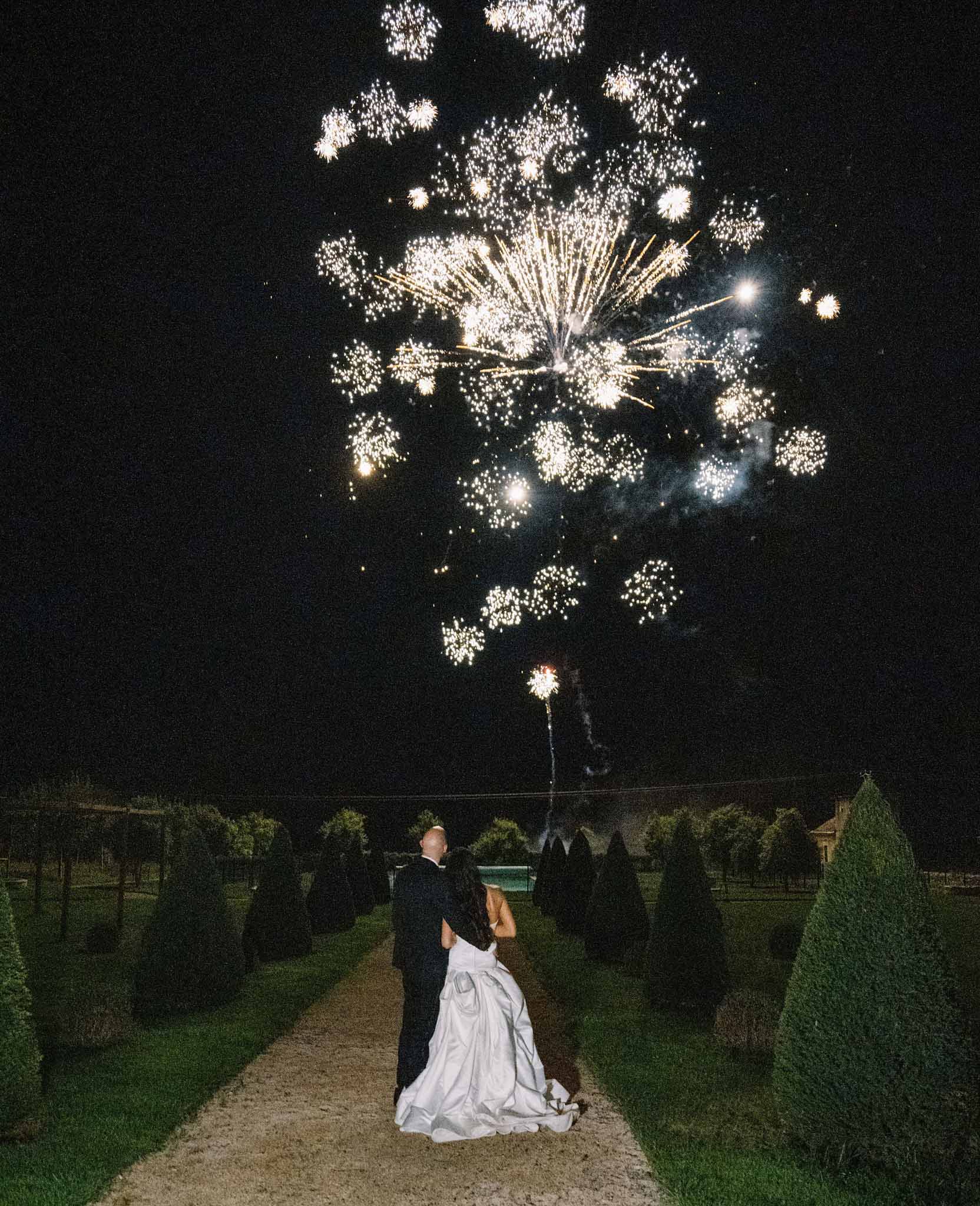 Bride and groom watching fireworks from a garden pathway lined with topiary hedges at night
