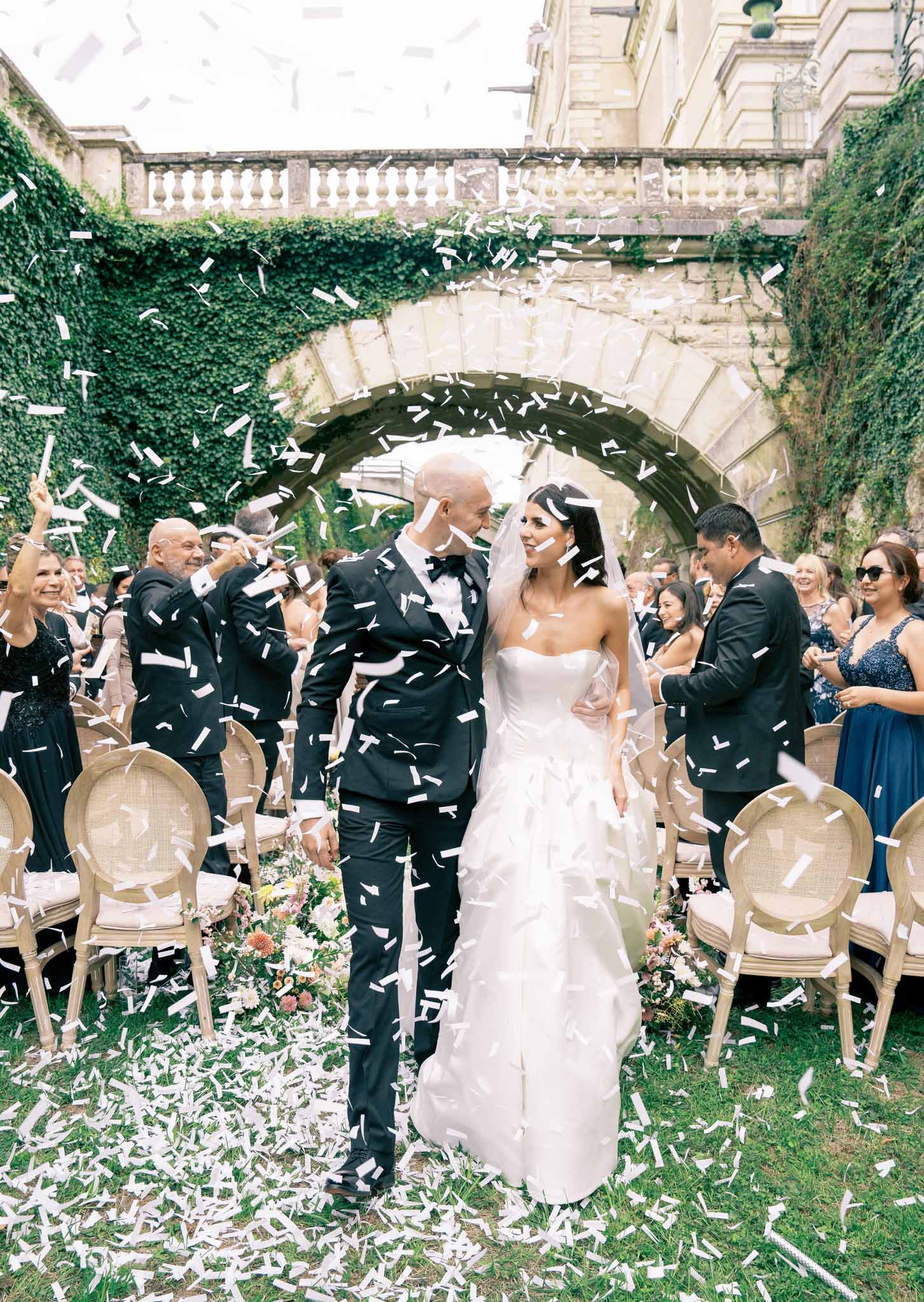 Bride and groom walk down aisle through white confetti in ivy-covered stone courtyard after ceremony