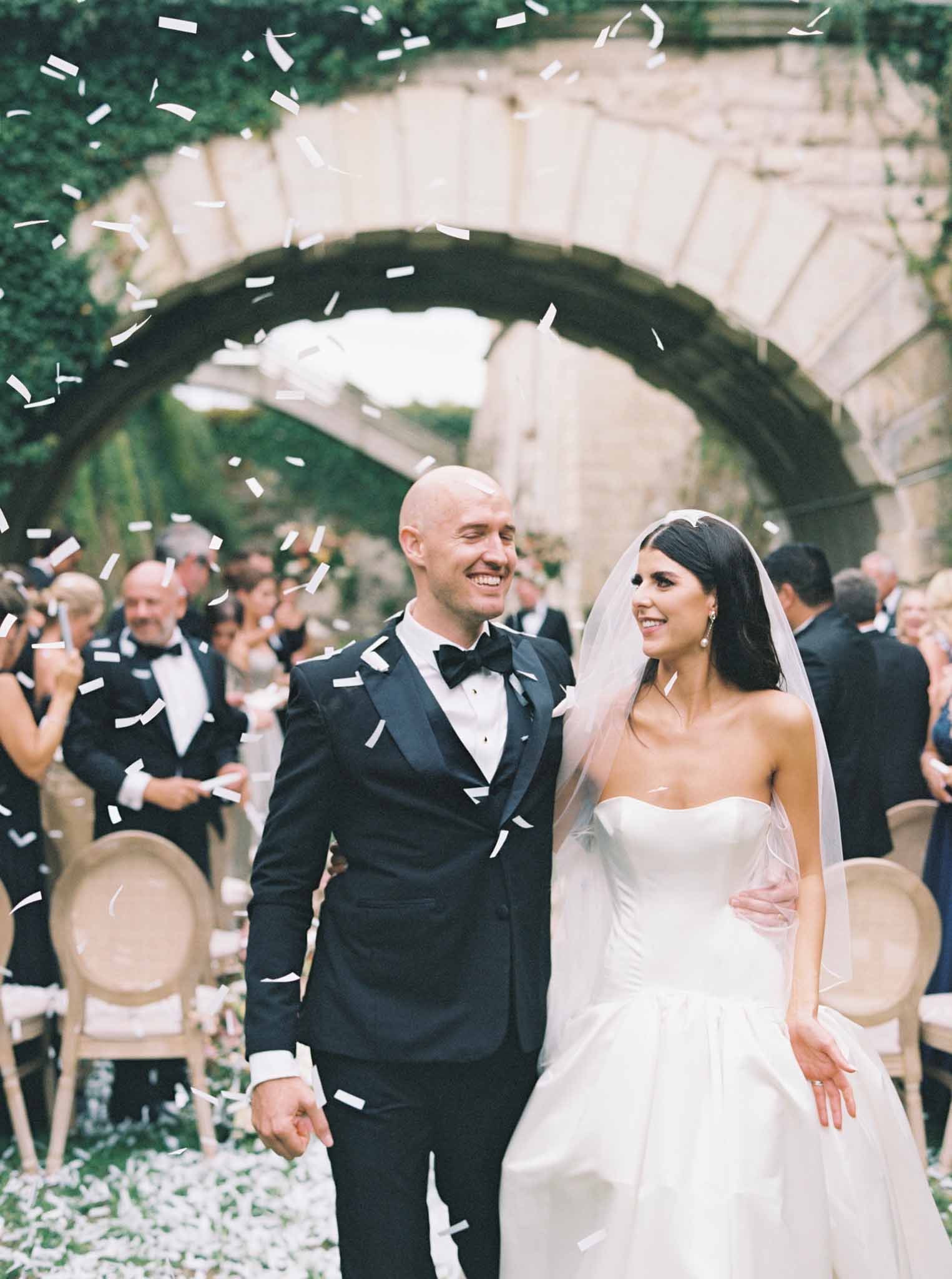 Bride and groom exit down the aisle as guests shower them with white confetti in an ivy-covered stone courtyard