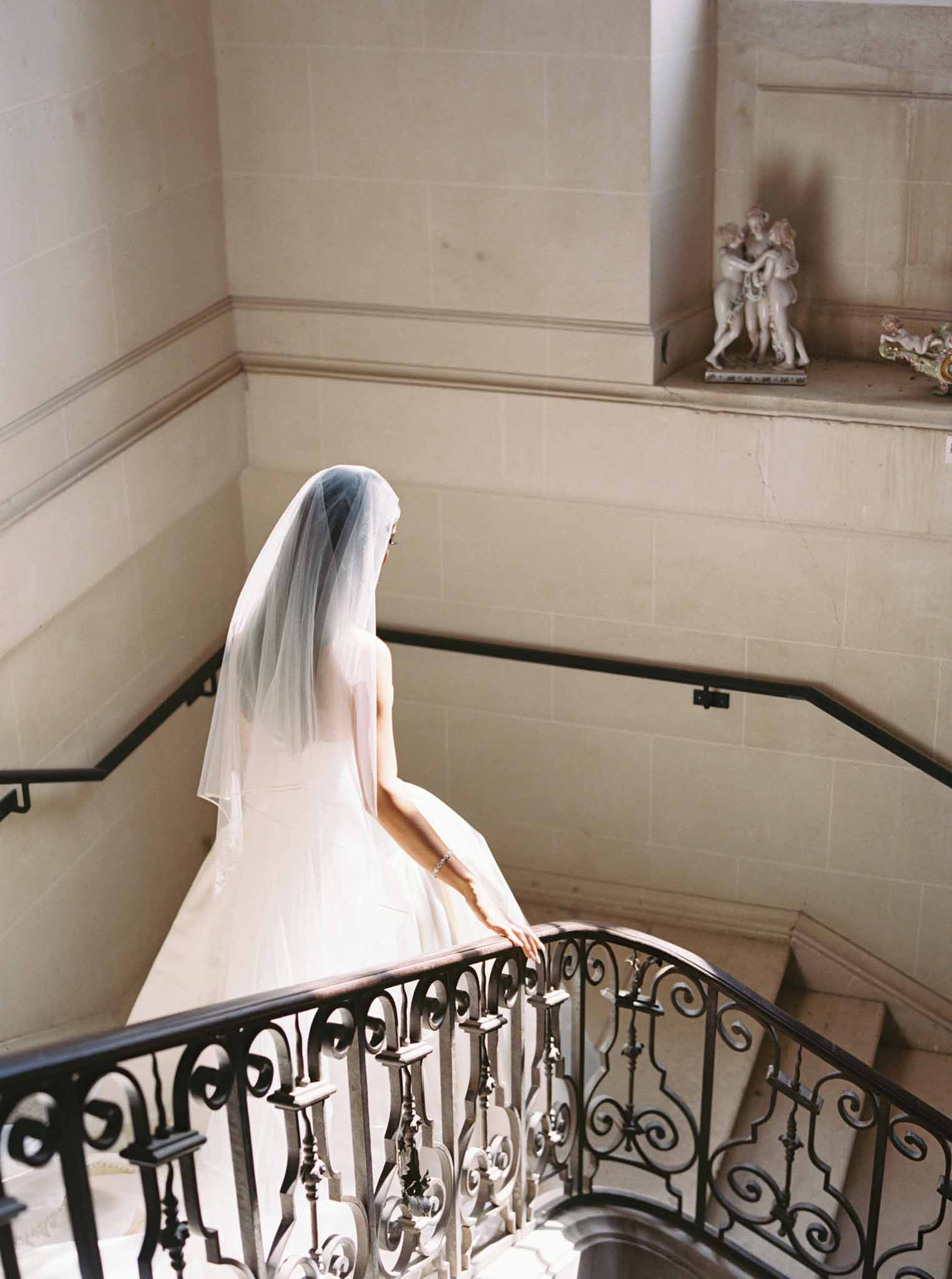 Bride from behind descending grand stone staircase with ivory tulle veil, black wrought-iron railing on walls