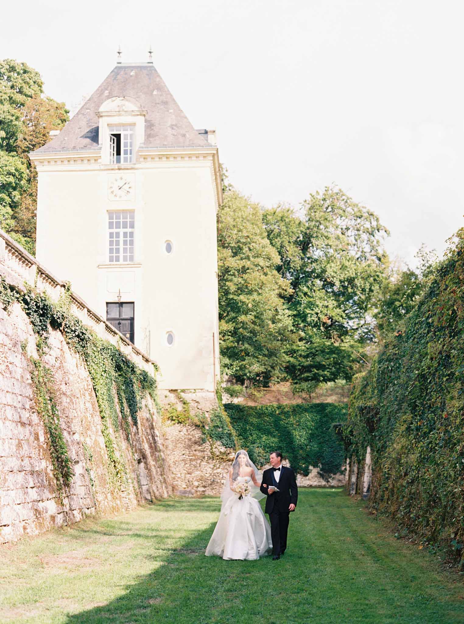 Bride and groom walking through walled garden toward ivy-covered chateau with mansard roof and clock face