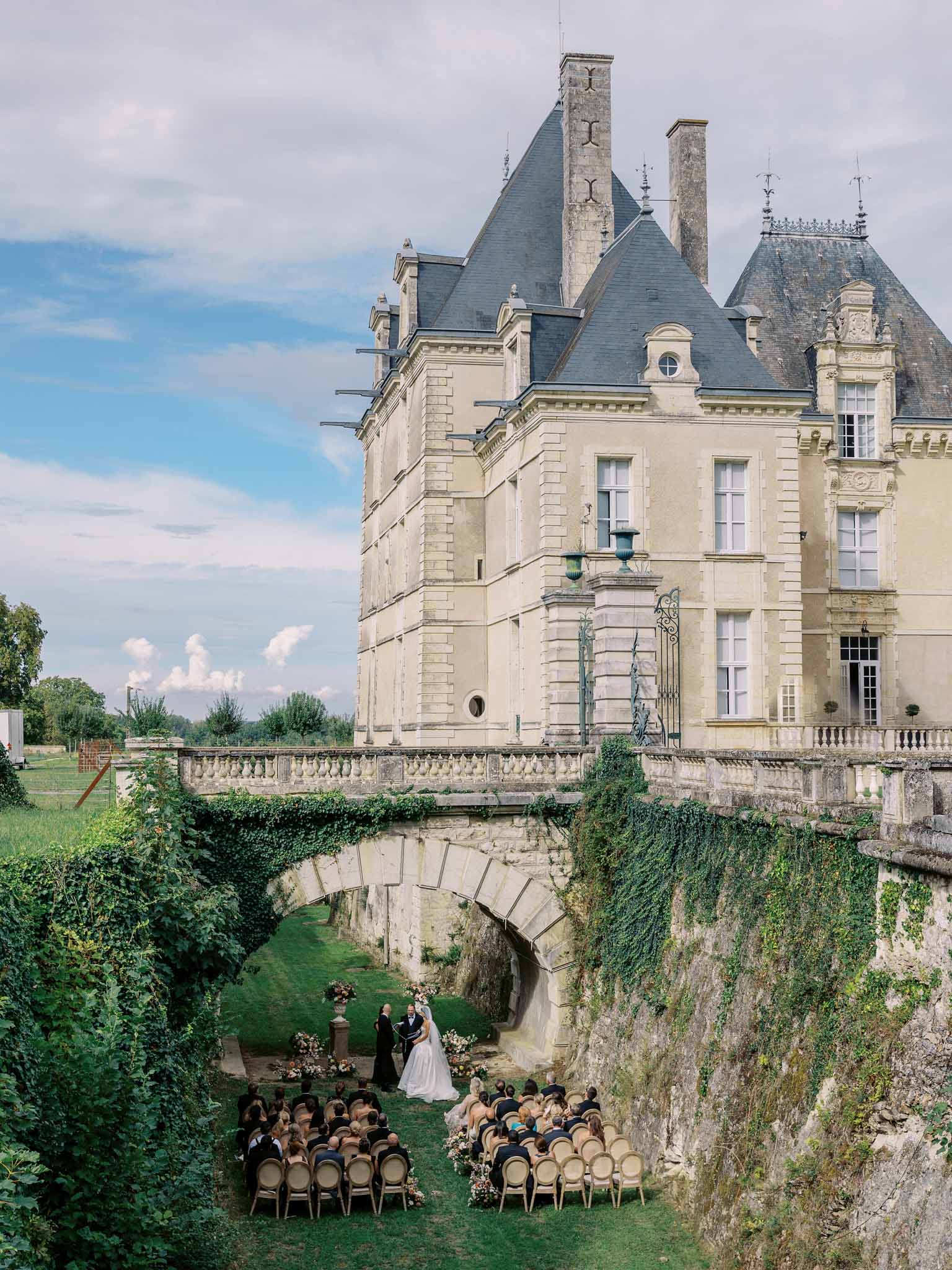 Wedding ceremony in château moat beneath stone bridge with guests seated facing couple and château rising behind