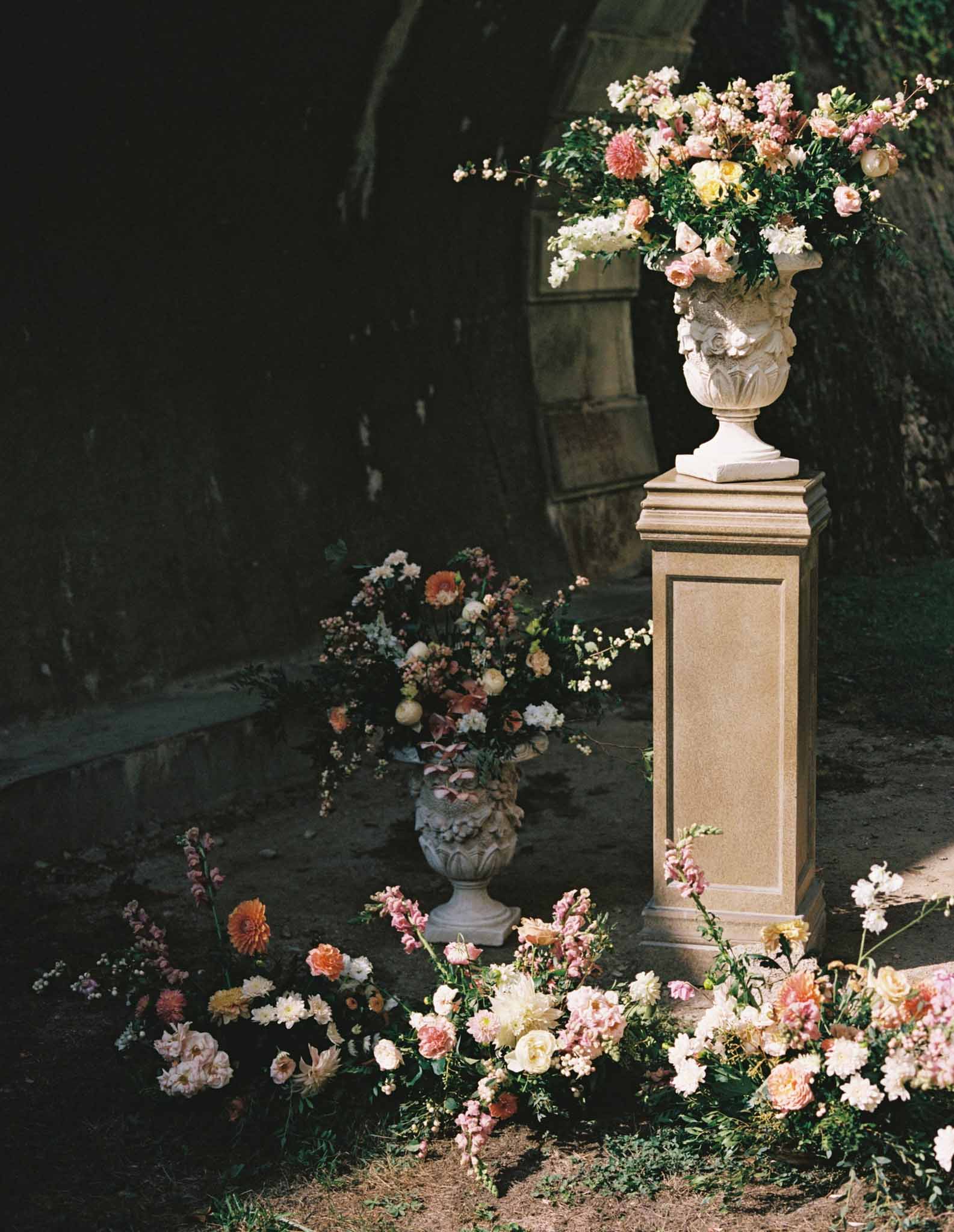 Two stone pedestals with ornamental urns of dusty rose, coral, and cream flowers in dark archway passage, loose blooms at base