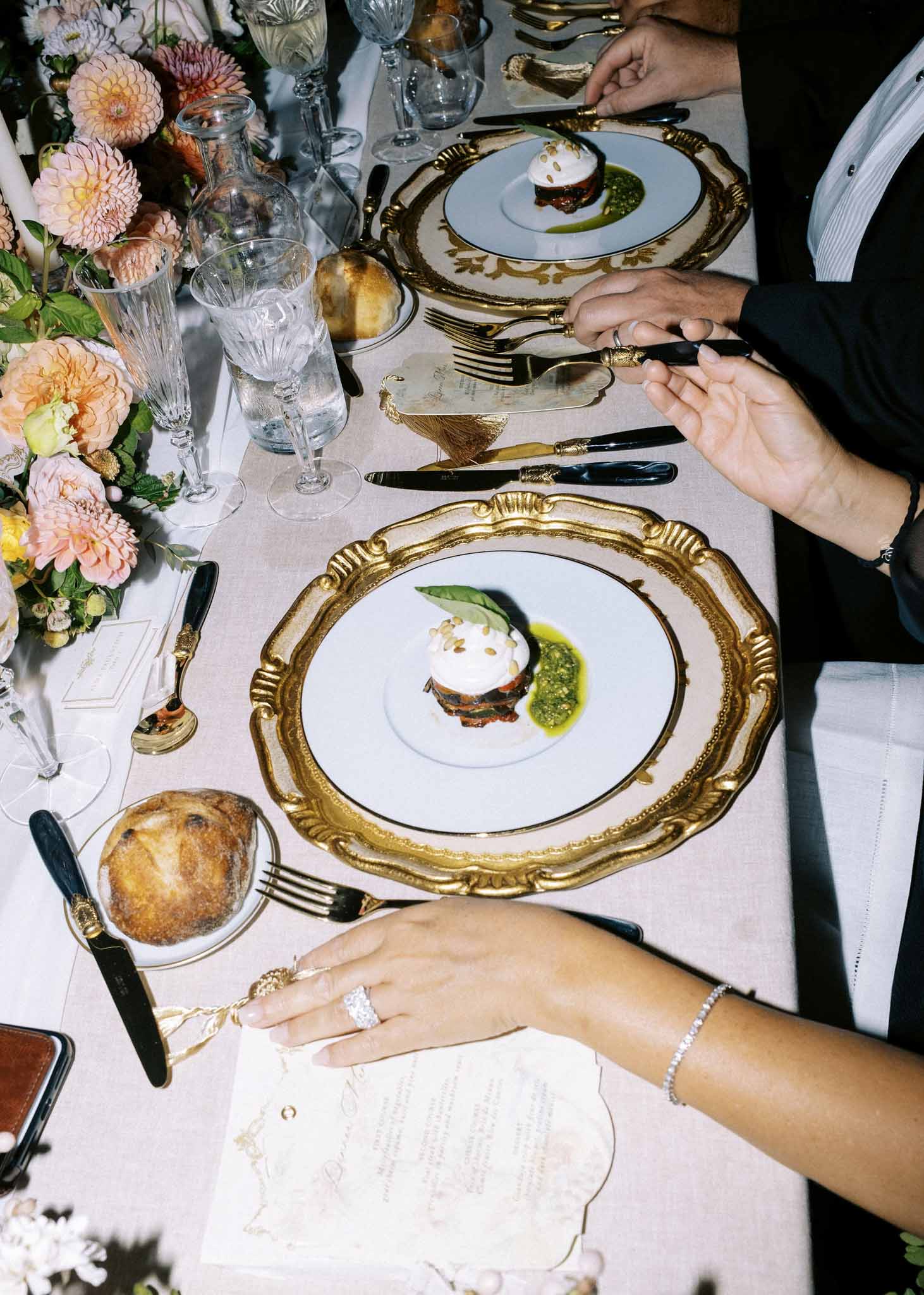 Close-up of plated entrée on gold-rimmed charger with pink dahlias centerpiece, bride's ring hand visible in foreground